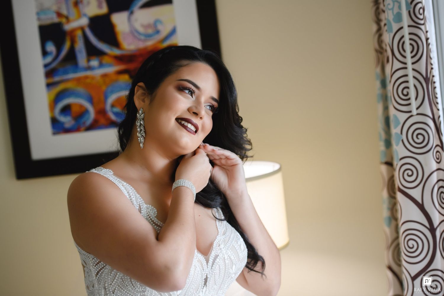 A bride in a white gown adjusts her earrings while smiling softly near a lamp and colorful artwork.