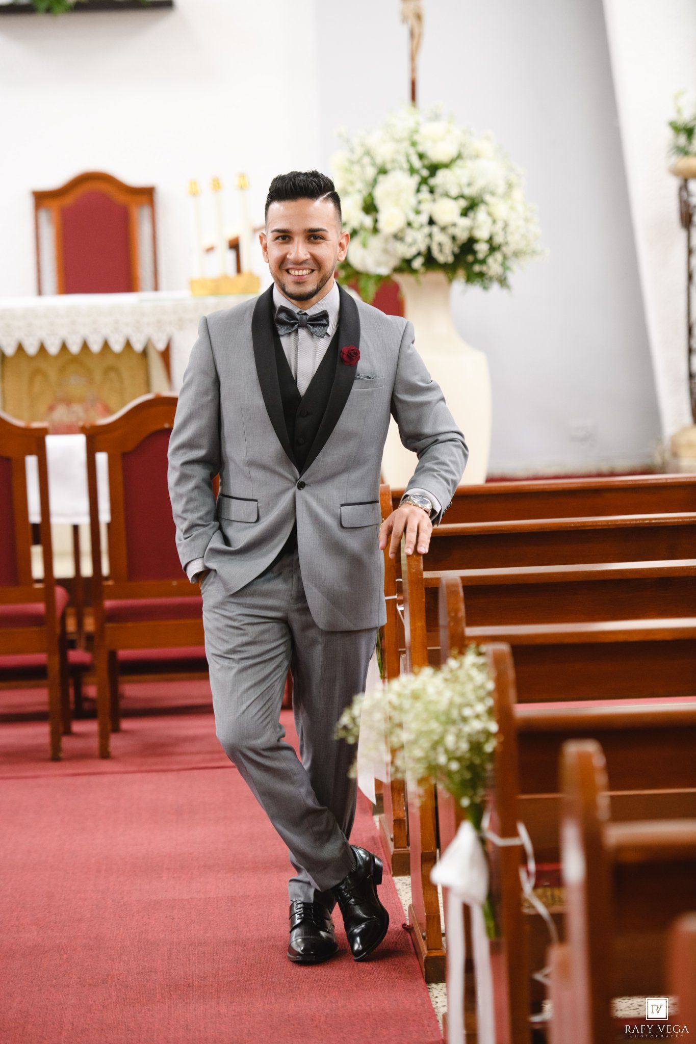 Groom in gray tuxedo smiling while leaning on church pew decorated with white flowers on red carpet aisle.