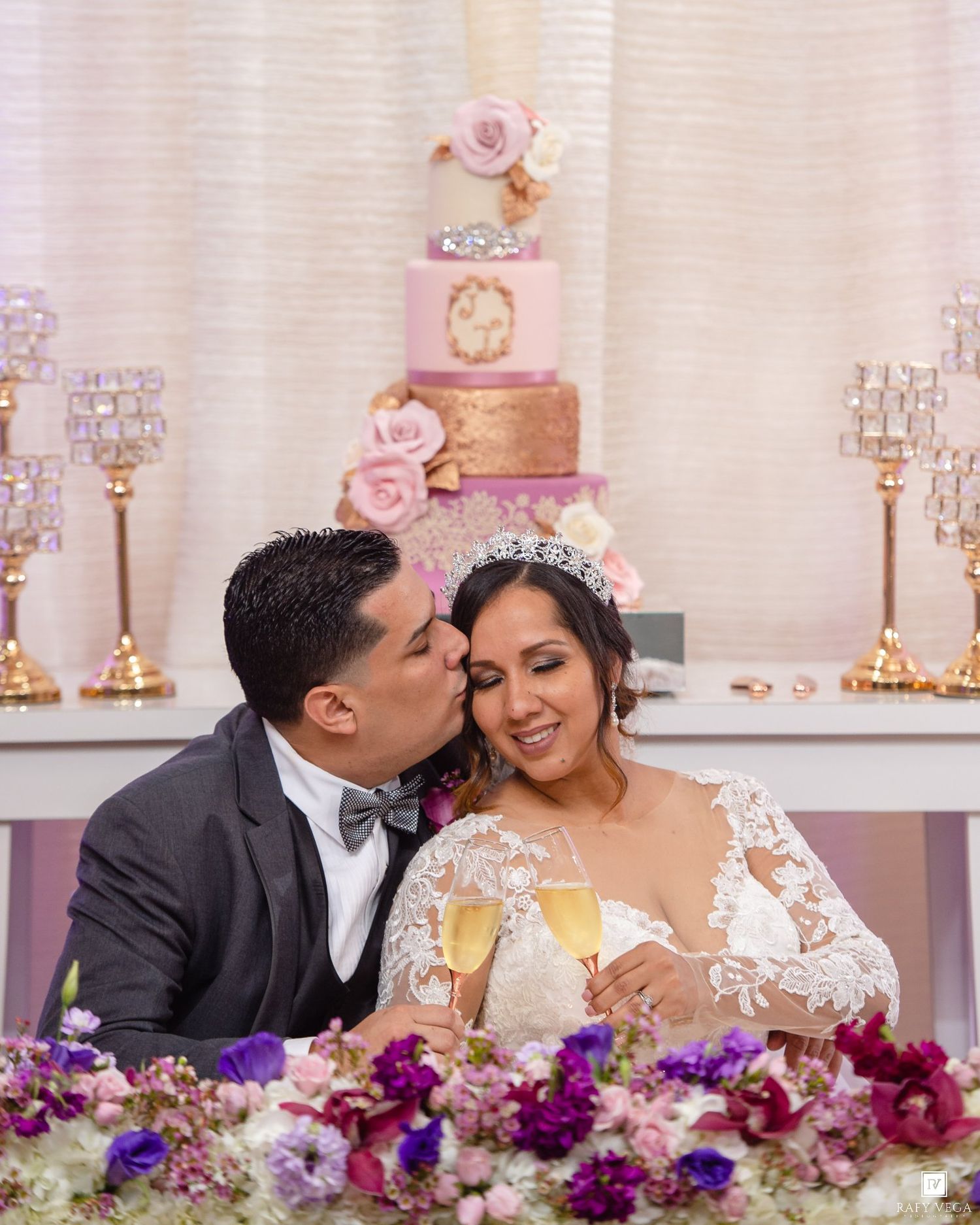 Bride and groom toast with champagne at wedding reception sweetheart table with pink floral cake backdrop.