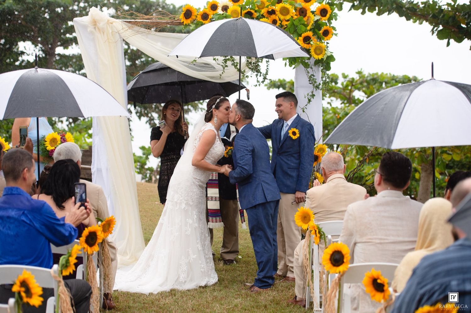 Bride and groom share a kiss at outdoor wedding ceremony decorated with sunflowers, guests holding umbrellas on a rainy day.