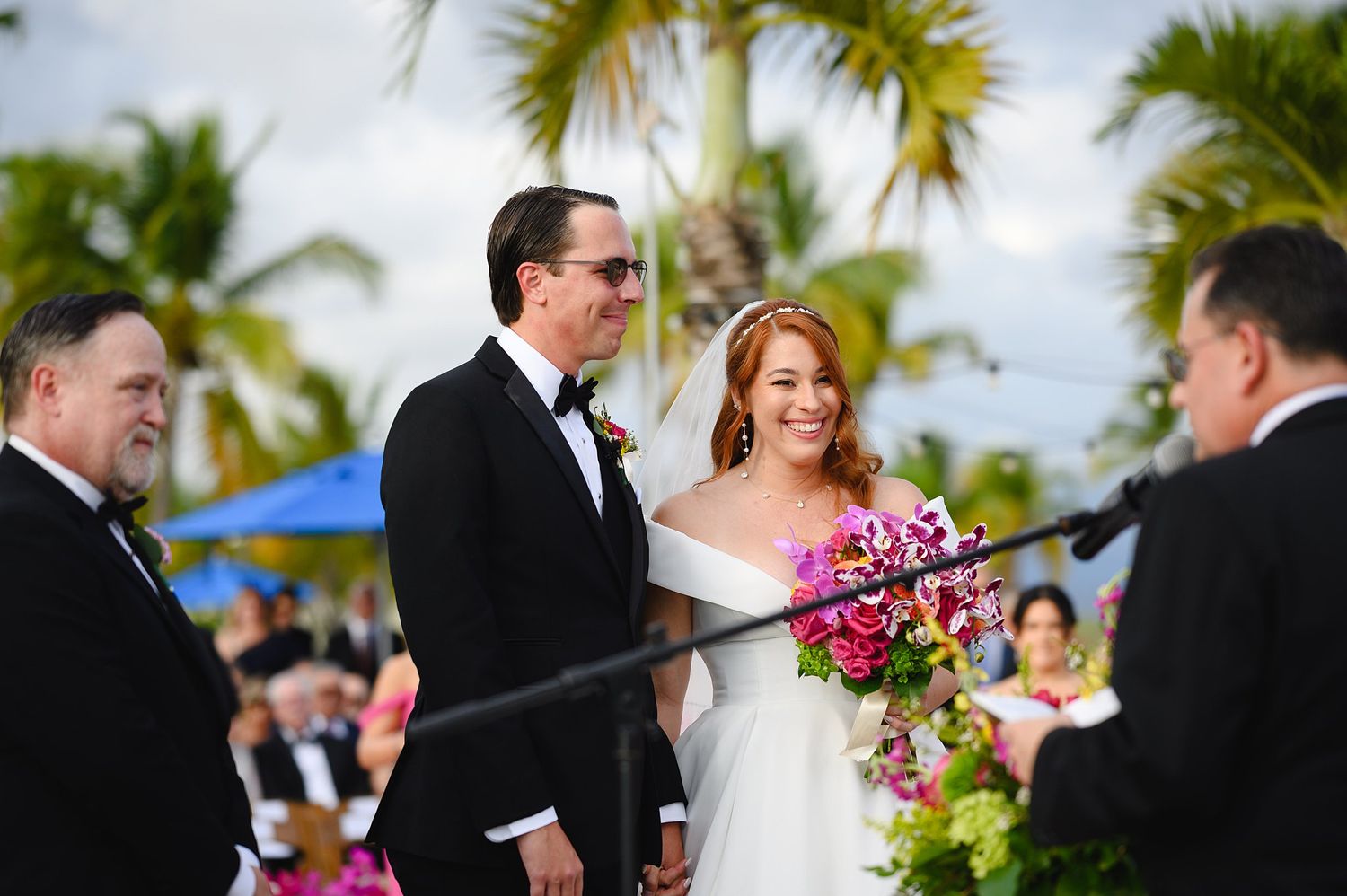 Bride and groom smiling during outdoor tropical wedding ceremony with lush palm trees and vibrant pink bouquet.