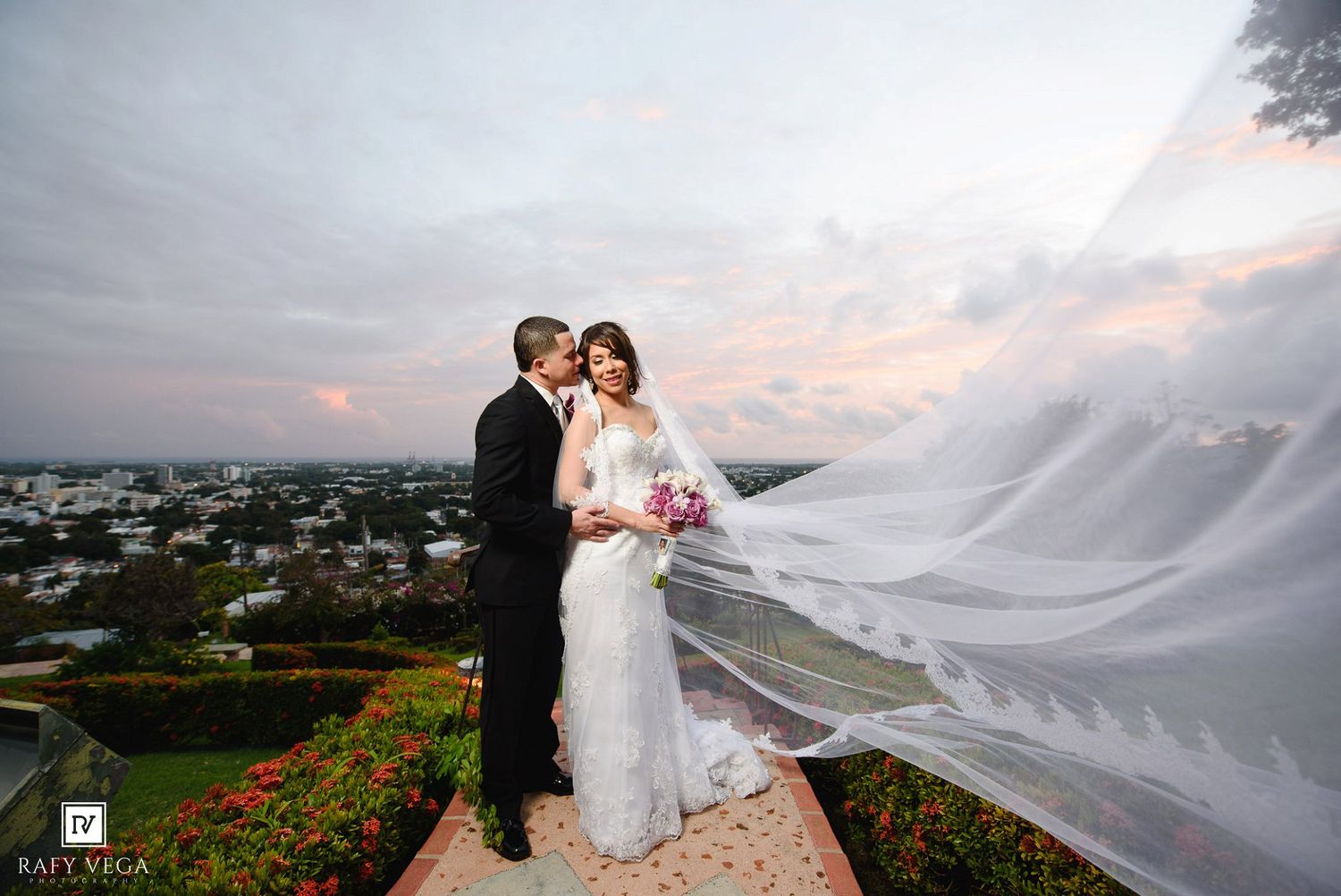 Bride and groom embrace at sunset rooftop venue, her veil flowing dramatically in the wind against a colorful sky.