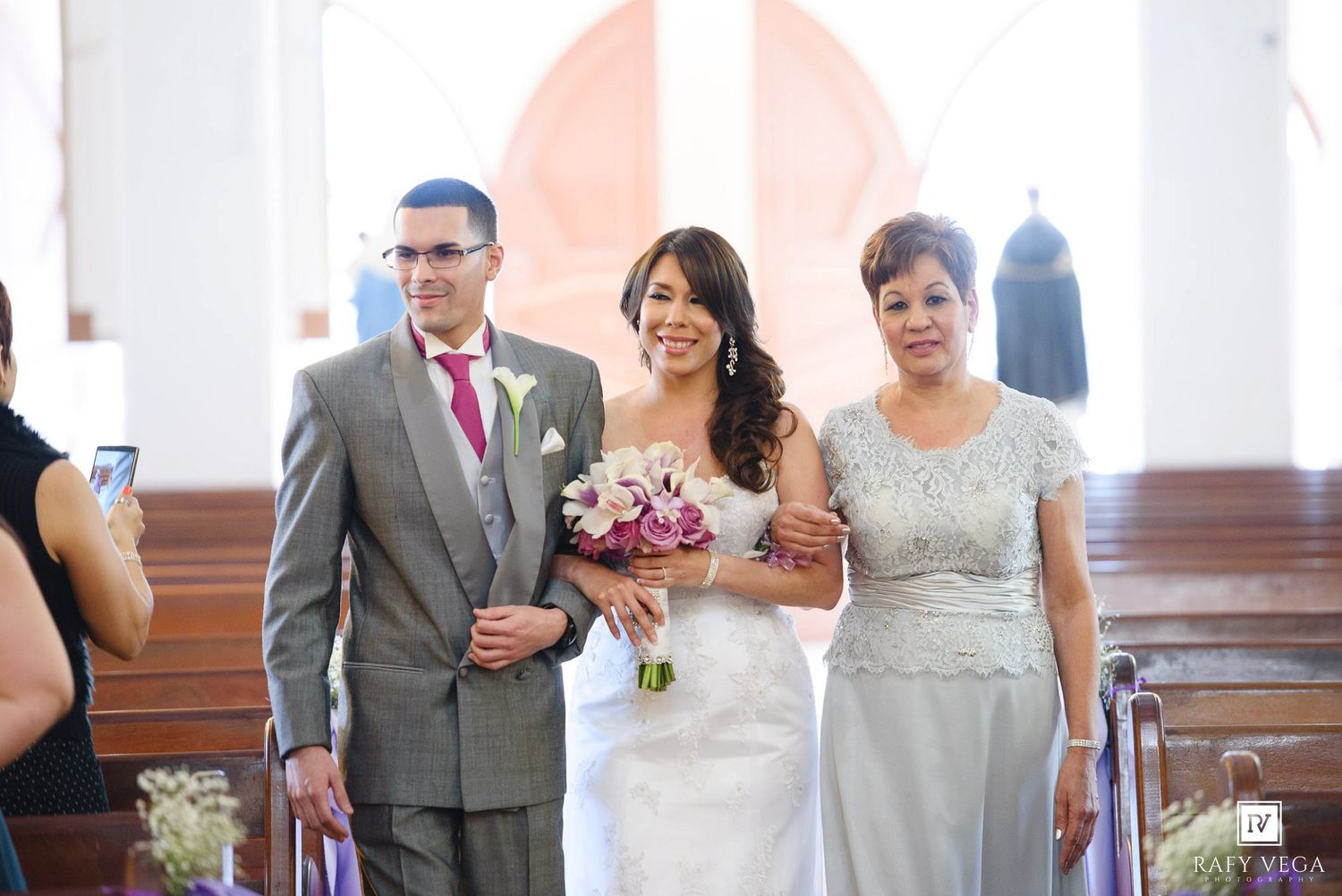 Bride walks down the aisle with parents in a sunlit church during a wedding ceremony.