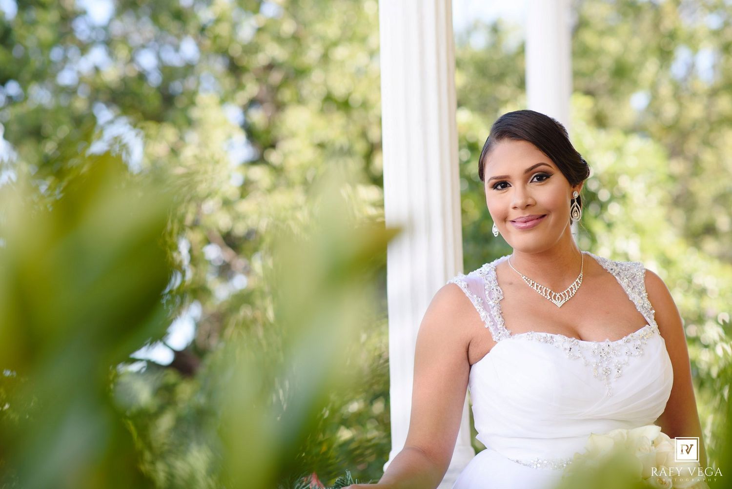 Smiling bride in white lace gown and diamond necklace poses gracefully between white columns surrounded by lush greenery.