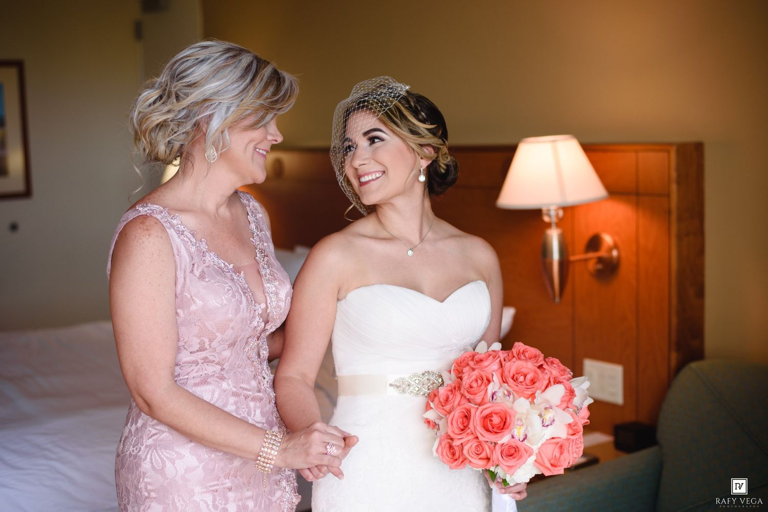Bride in white strapless gown holds pink bouquet beside woman in pink lace dress in elegant hotel room.