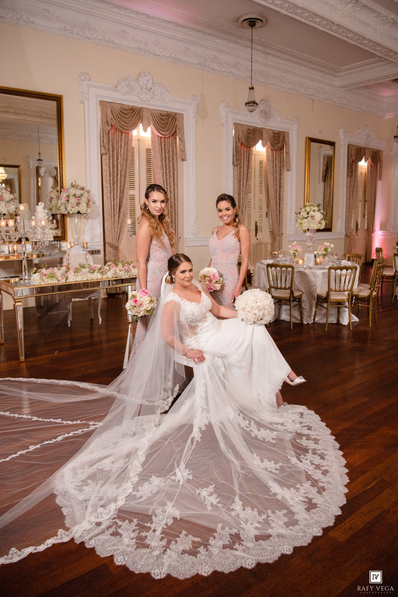 Bride in white gown with lace veil poses with two bridesmaids in pink dresses in elegant ballroom reception.