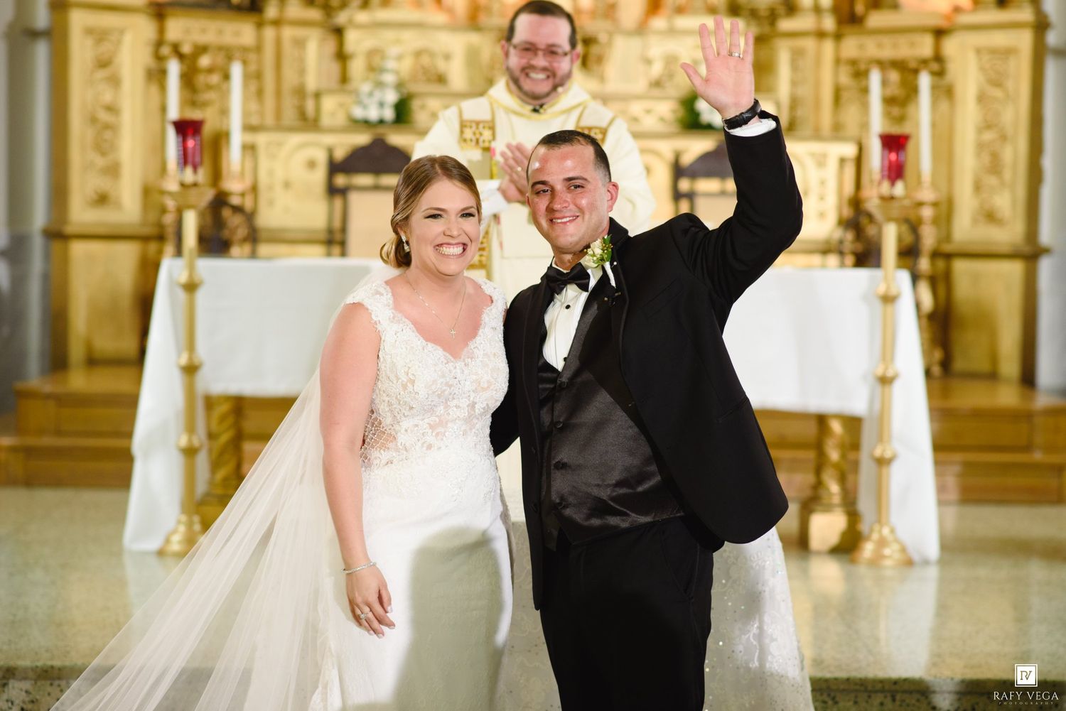 Smiling newlywed couple poses at church altar with priest behind them during wedding ceremony.