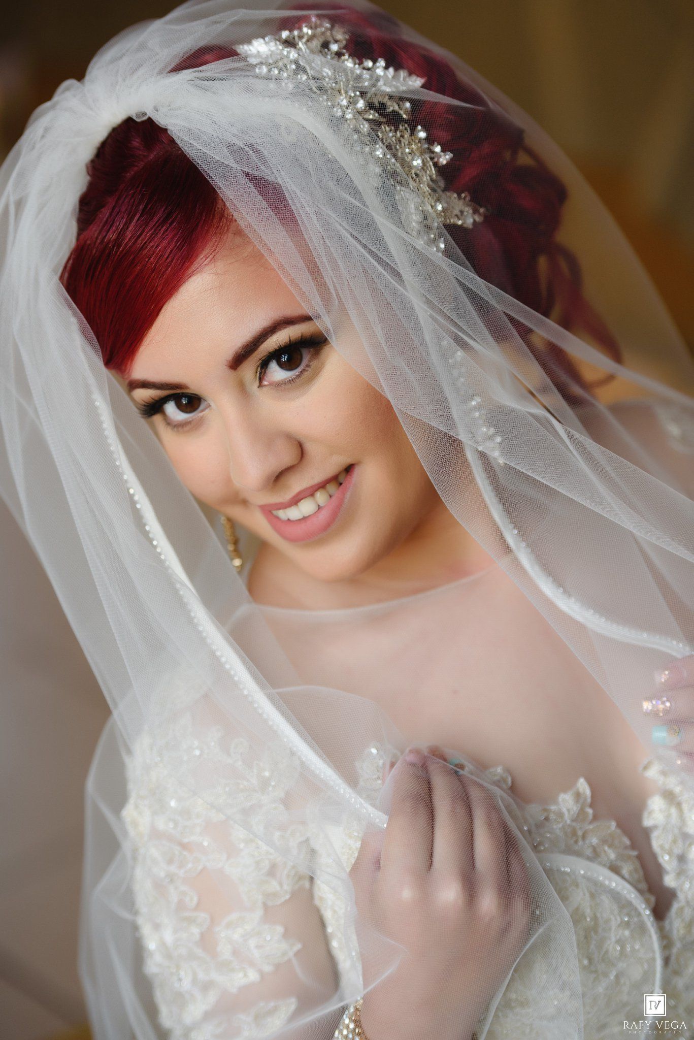 Smiling bride with red hair wearing a white lace wedding dress and veil adorned with a crystal hair piece.