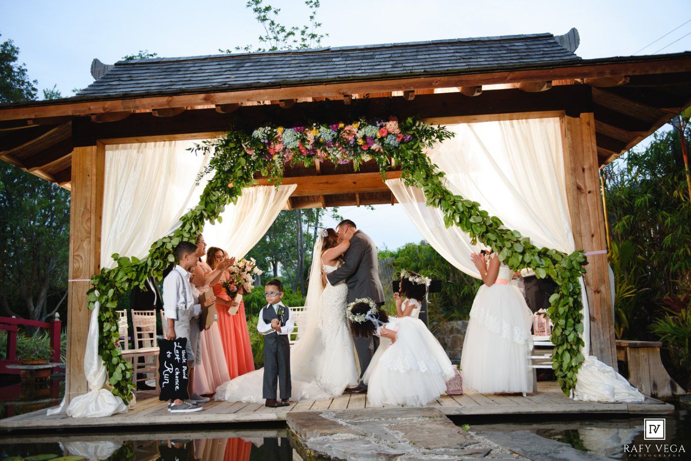 Romantic outdoor wedding ceremony under a floral arch with bride and groom kissing, surrounded by bridesmaids and flower girls.