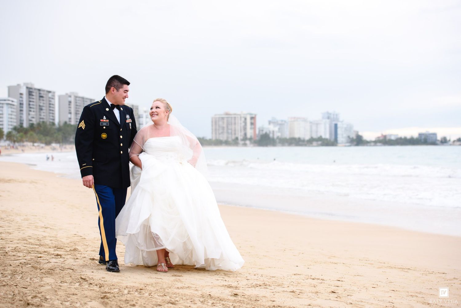 Military groom in dress uniform walks with bride in white gown on sandy beach with city skyline backdrop.
