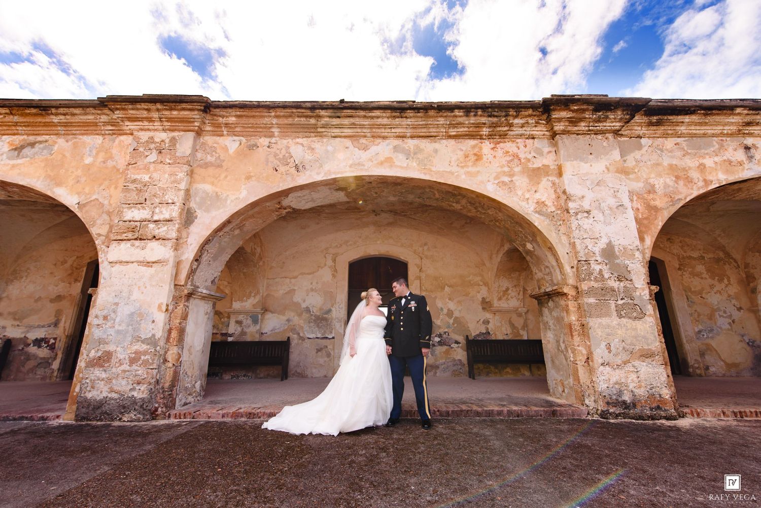 Bride and groom share a romantic moment under stone arches at a historic venue with blue skies.