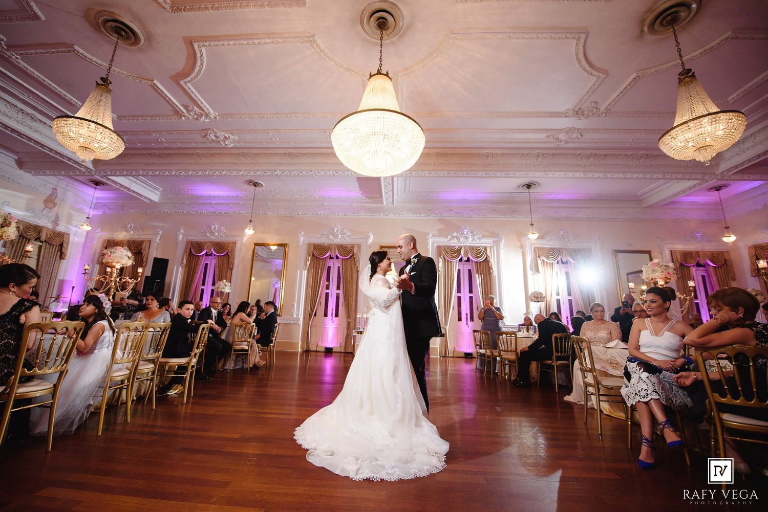 Bride and groom share first dance in elegant ballroom with chandeliers and purple uplighting at wedding reception.