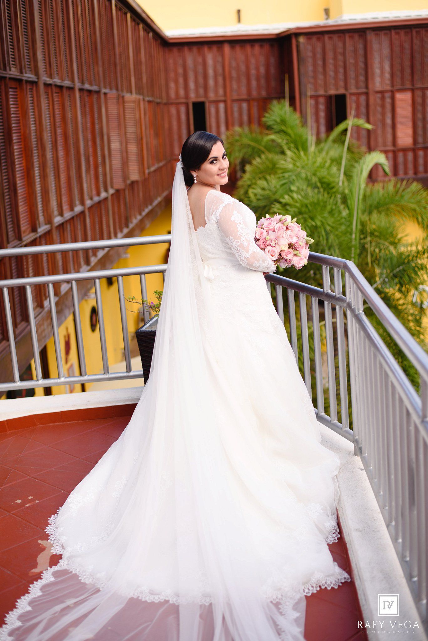 Bride in white lace long-sleeve gown with veil and pink bouquet poses on balcony at outdoor wedding venue.