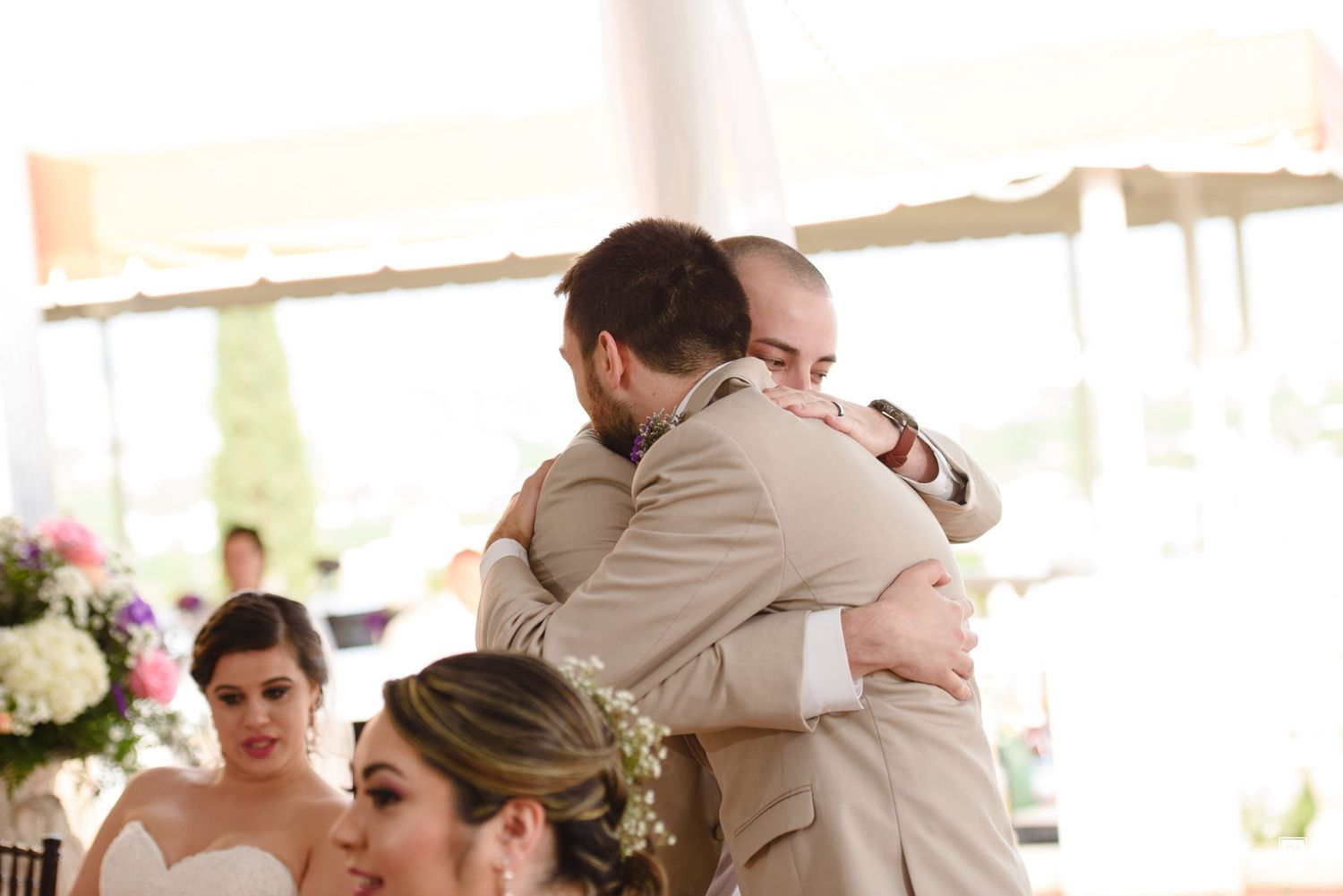 Two men share an emotional hug at a wedding reception while the bride watches nearby.