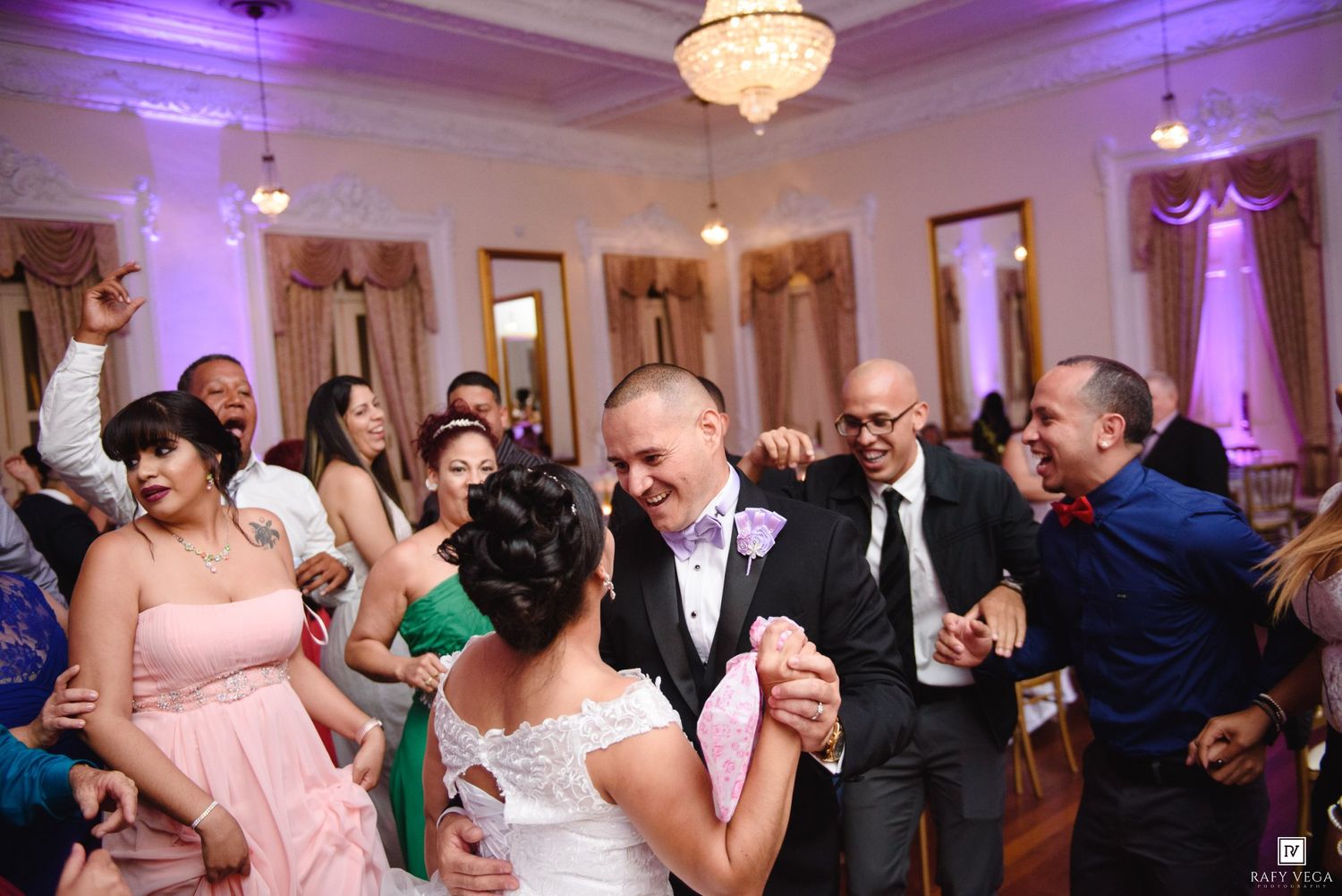 Bride and groom dancing joyfully with guests at elegant wedding reception in decorated ballroom.