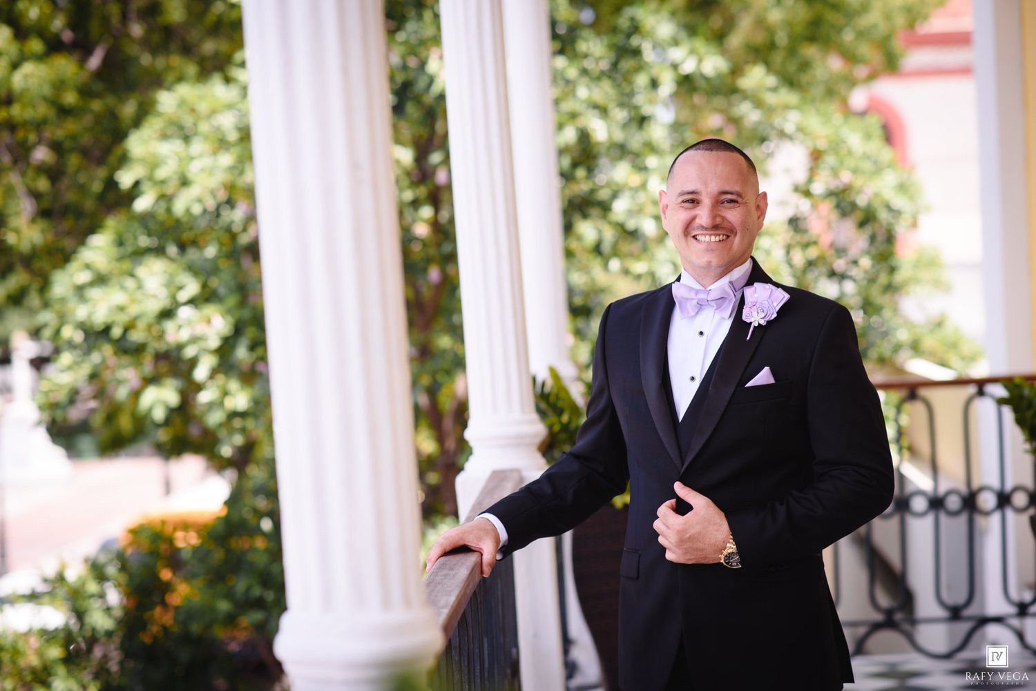 Smiling groom in black tuxedo with lavender bow tie and boutonniere poses on elegant white columned venue porch.
