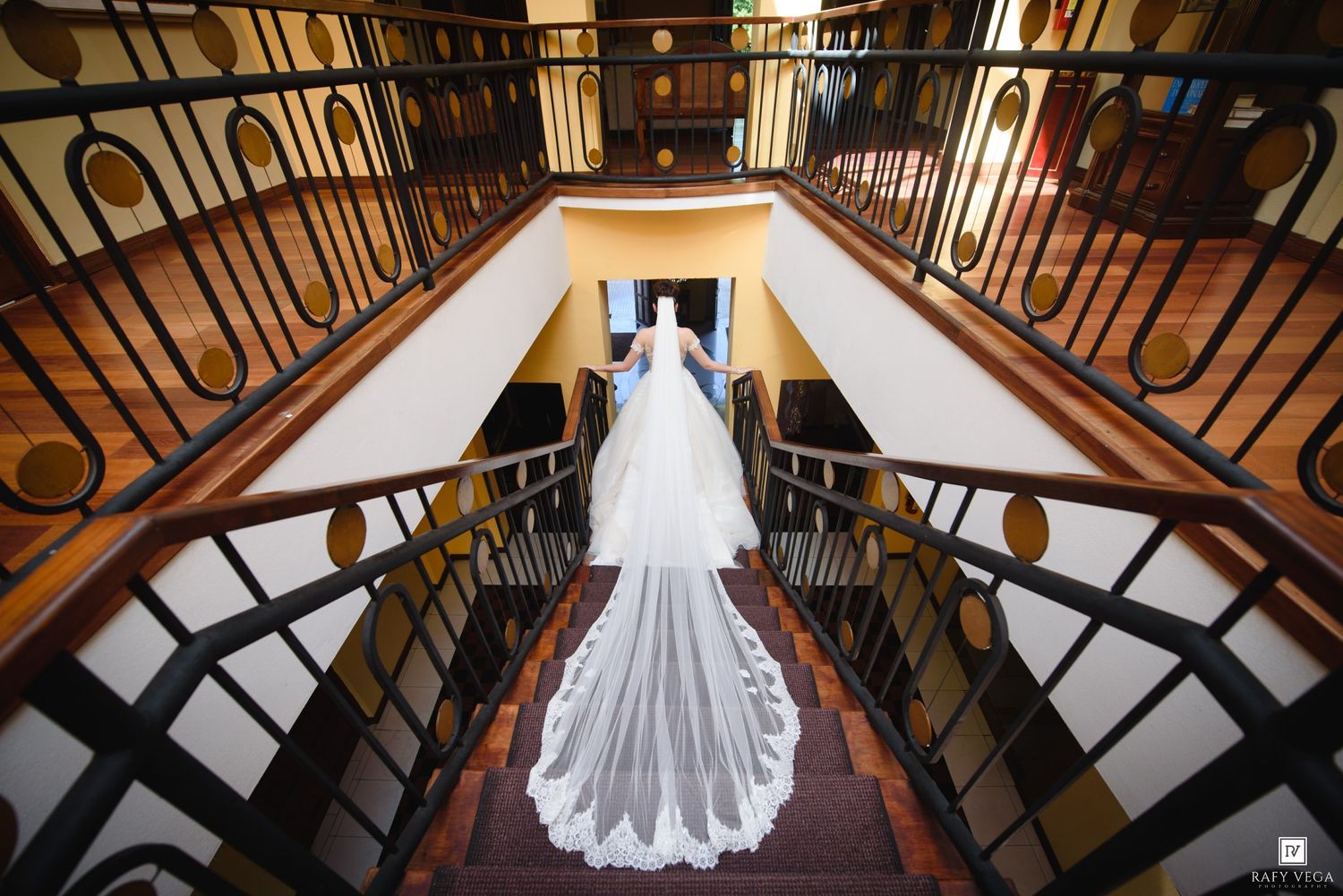 Bride in white gown with long veil stands atop elegant staircase in grand venue, captured from above.