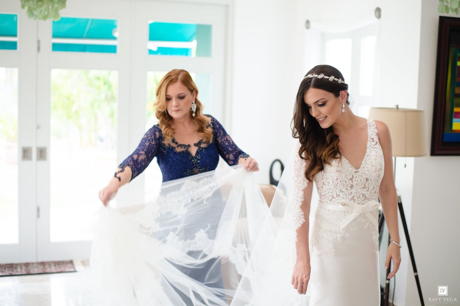Bride in white gown with bridesmaid in blue lace dress arranging wedding veil indoors before ceremony.