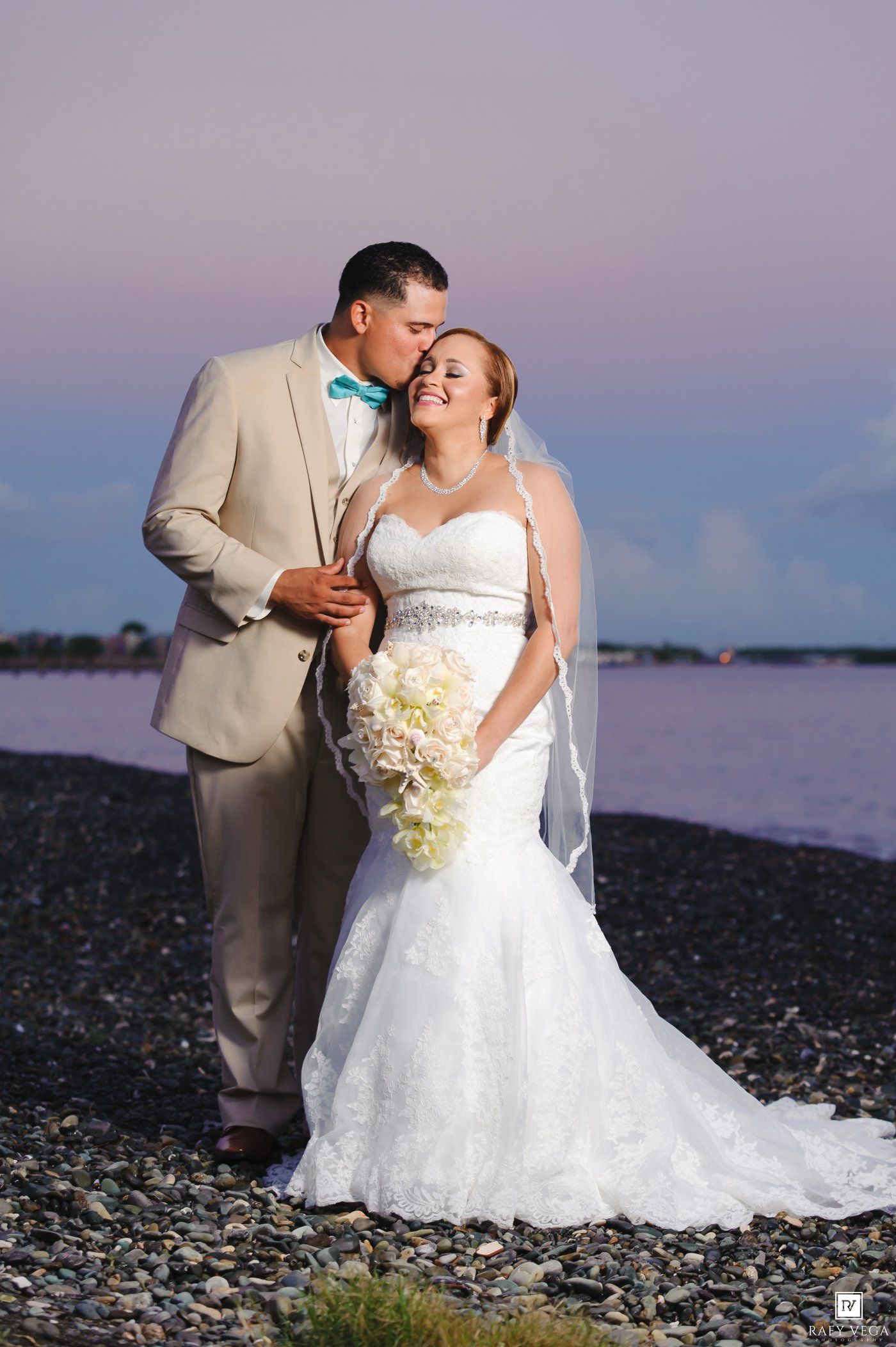 Bride and groom share a romantic kiss on a rocky beach at sunset, bride in mermaid gown holding white bouquet.