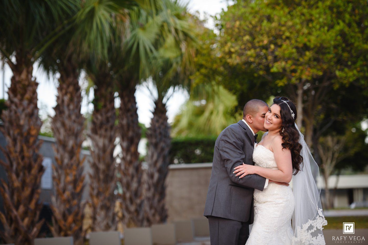 A bride and groom share a romantic kiss outdoors surrounded by lush palm trees on their wedding day.