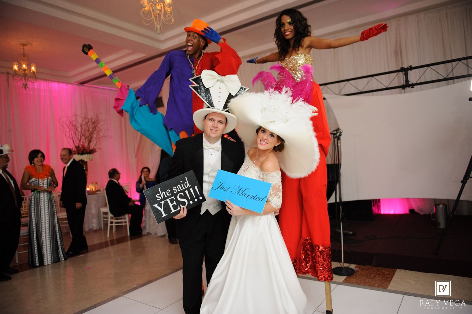 Bride and groom pose with colorful stilt walkers at elegant wedding reception holding celebratory signs.