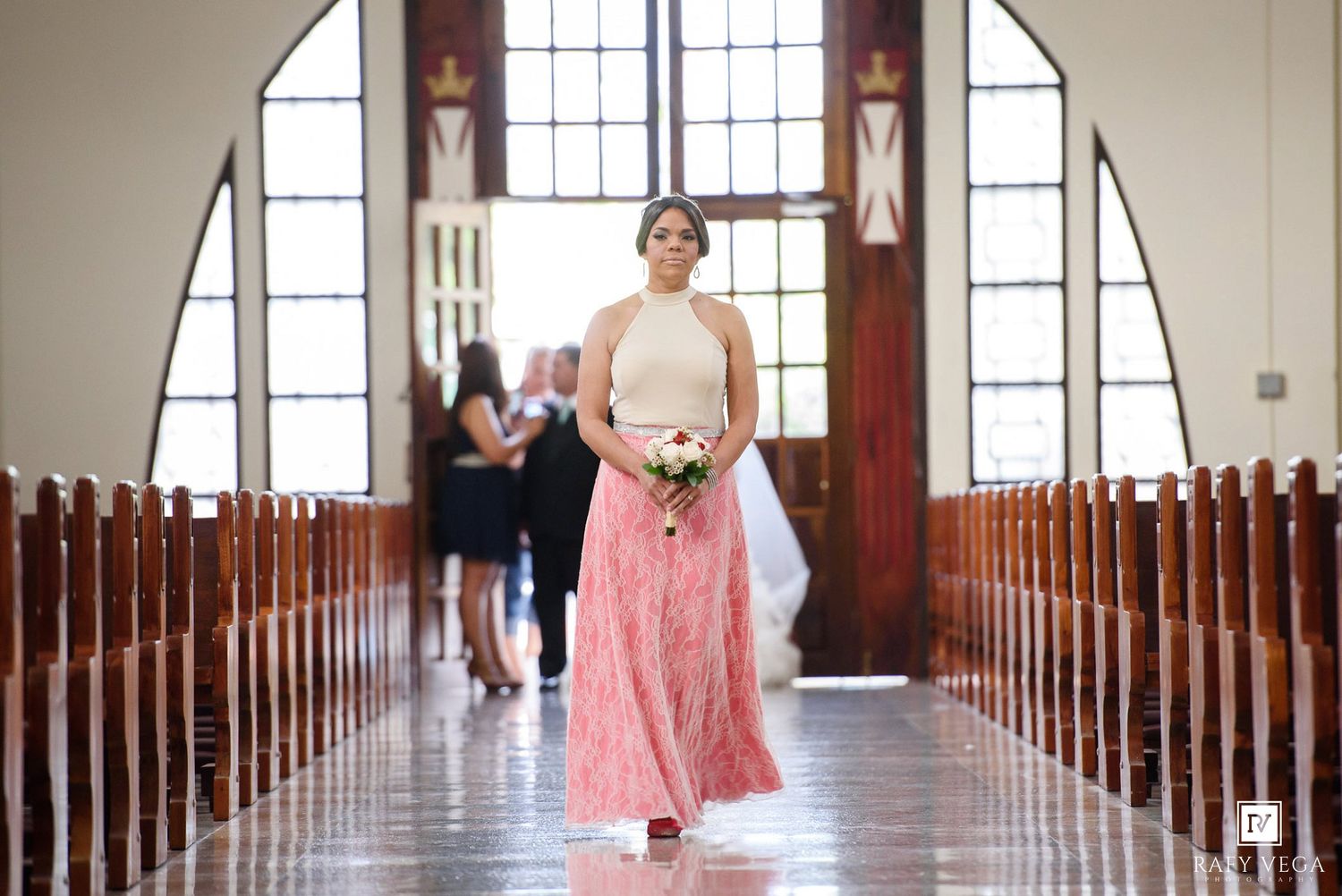 Bridesmaid in white top and pink lace skirt walks down church aisle holding bouquet during wedding ceremony.