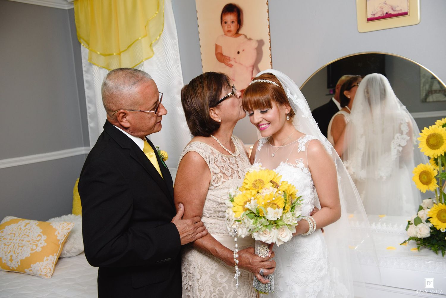 Bride in white gown receiving kiss from mother while father stands beside her before wedding.