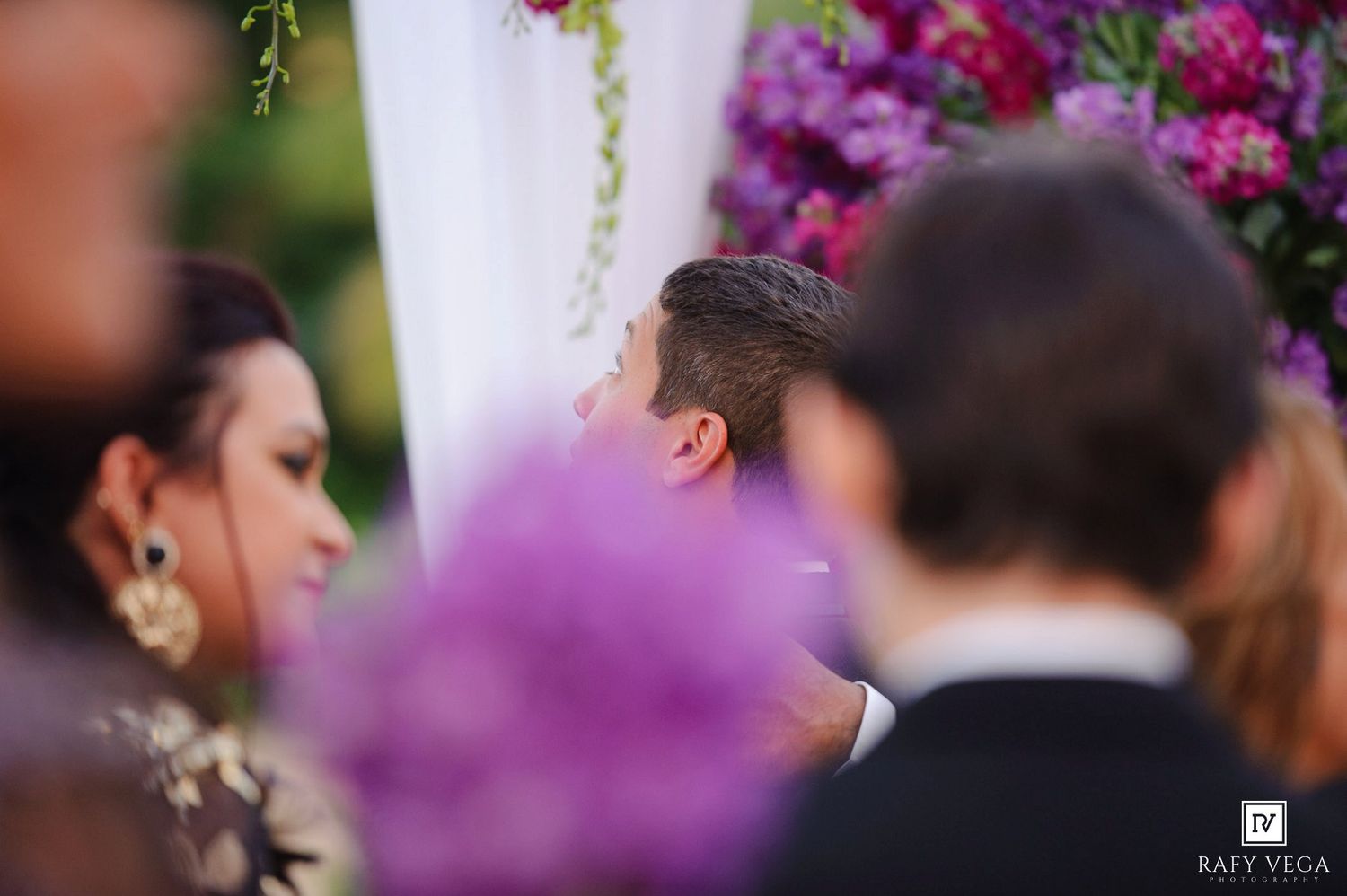 Guests gathered at an outdoor wedding ceremony with lush purple floral arrangements and greenery backdrop.