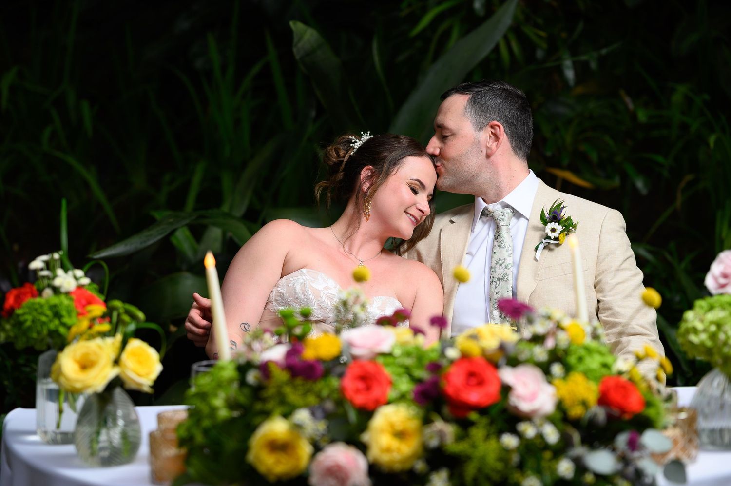 Bride and groom share a tender kiss at their floral-adorned wedding reception table surrounded by colorful blooms.