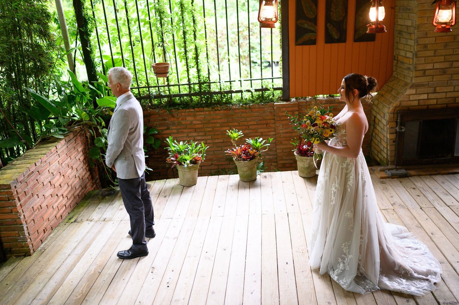 Bride in white gown holds bouquet facing groom on wooden deck surrounded by lush greenery and brick walls.
