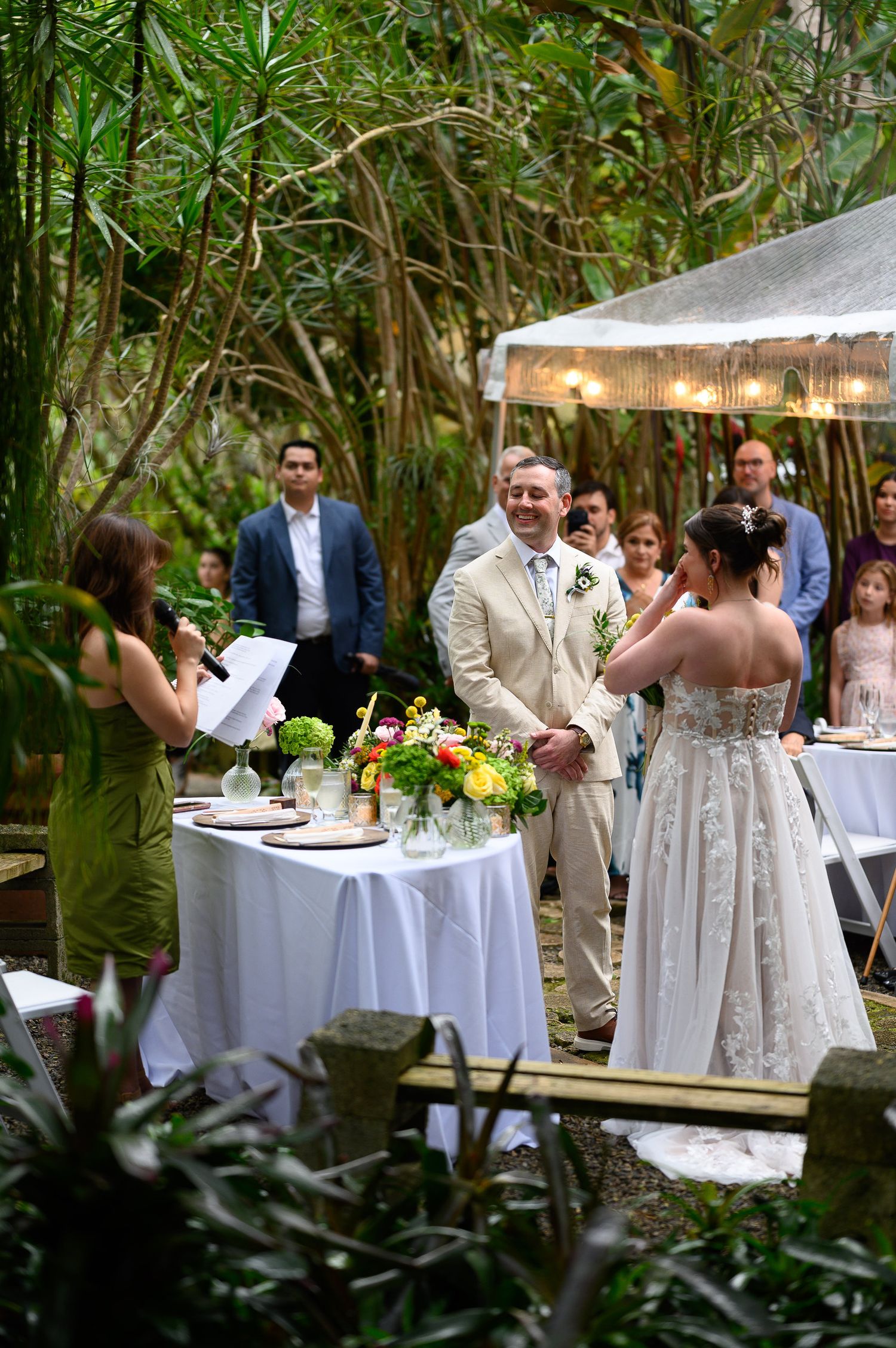 Outdoor garden wedding ceremony with bride and groom exchanging vows, officiant reading, guests seated among lush greenery.