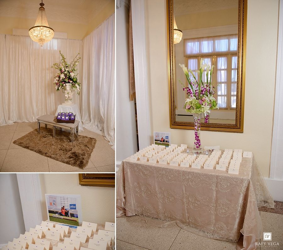 Elegant wedding venue escort card table with floral arrangements, candles, and a gold mirror in a white-draped reception hall.