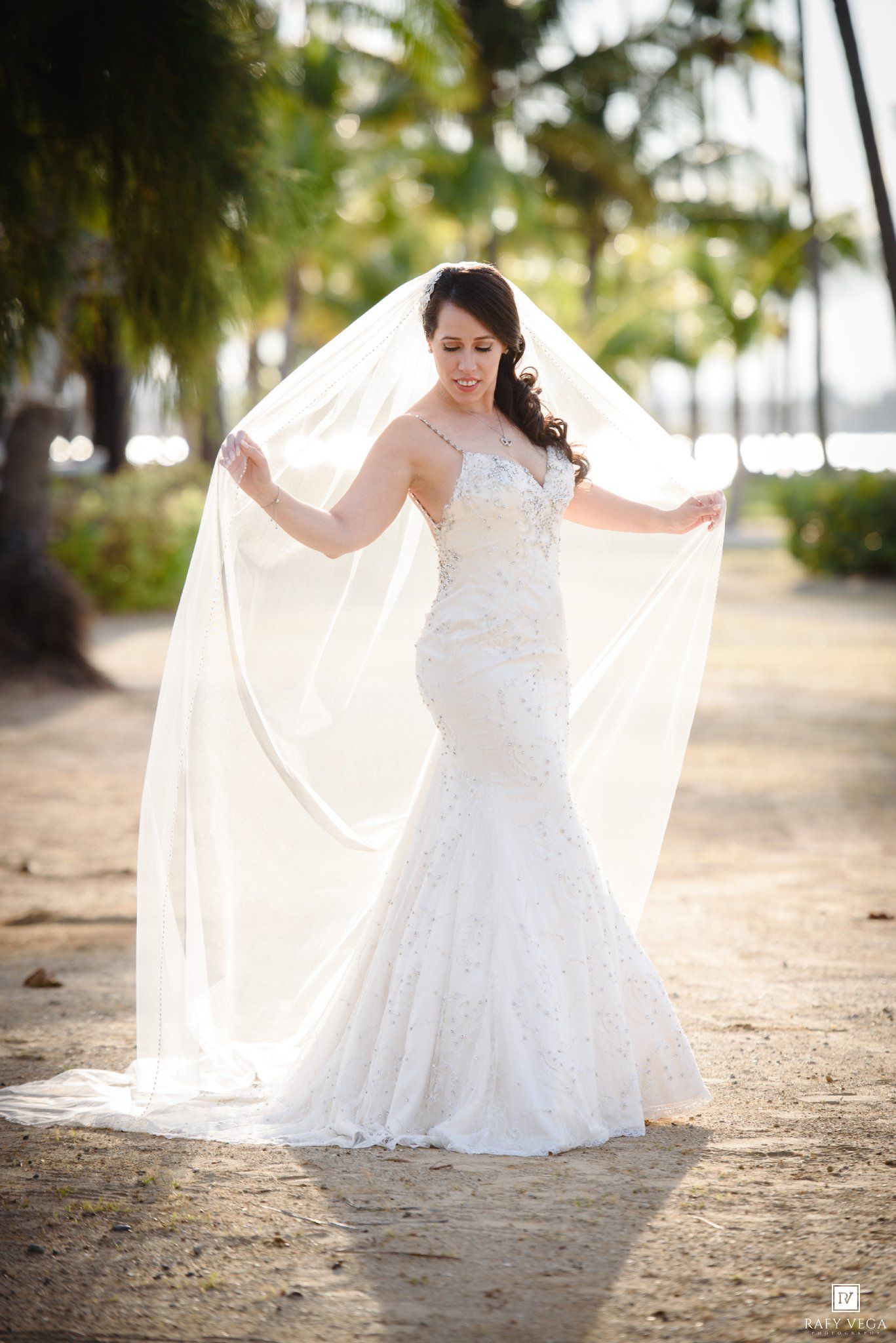 Bride in beaded mermaid gown and flowing veil poses outdoors among palm trees in a tropical wedding setting.