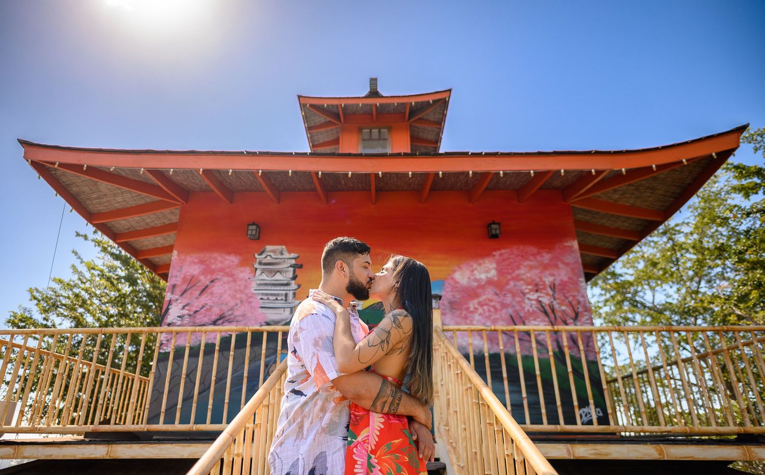 Couple sharing a romantic kiss on steps before a vibrant red Japanese pagoda with cherry blossoms on a sunny day.