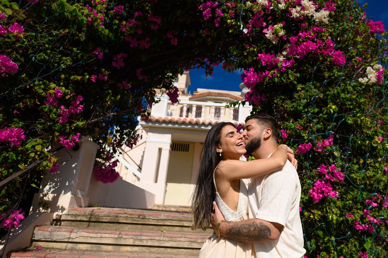 Couple embracing under vibrant pink bougainvillea flowers near a white building on sunny day.
