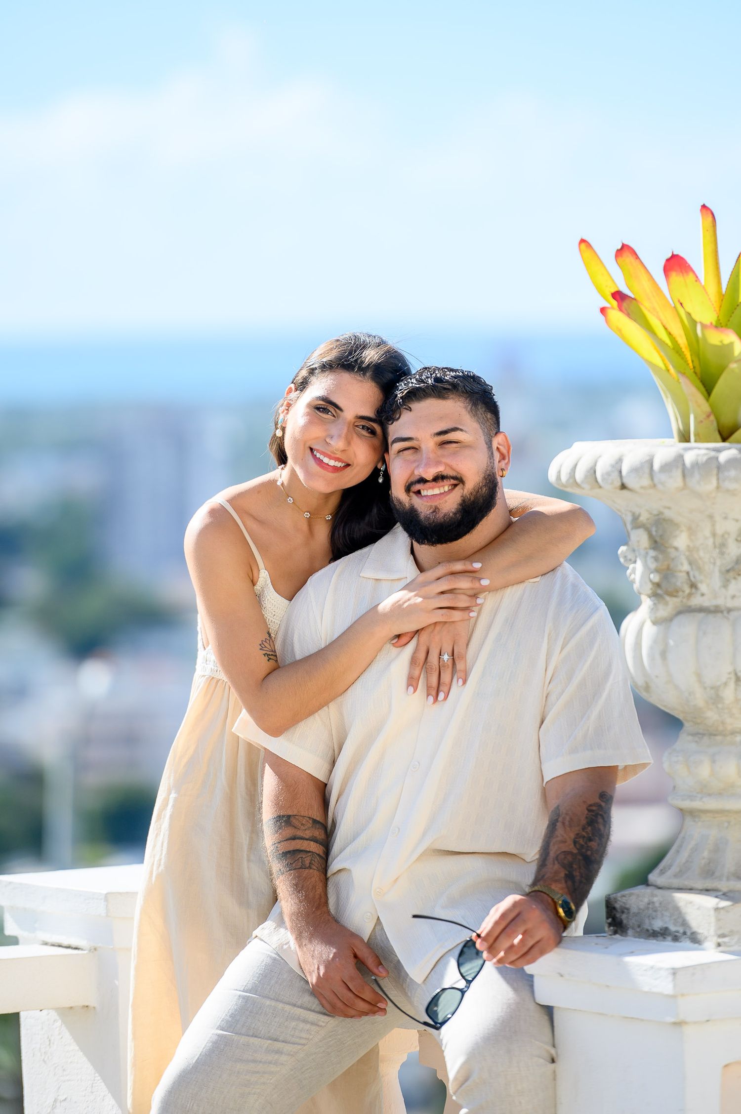 Smiling couple posing on a sunny terrace, woman in cream dress embracing tattooed man in beige outfit with city skyline behind.