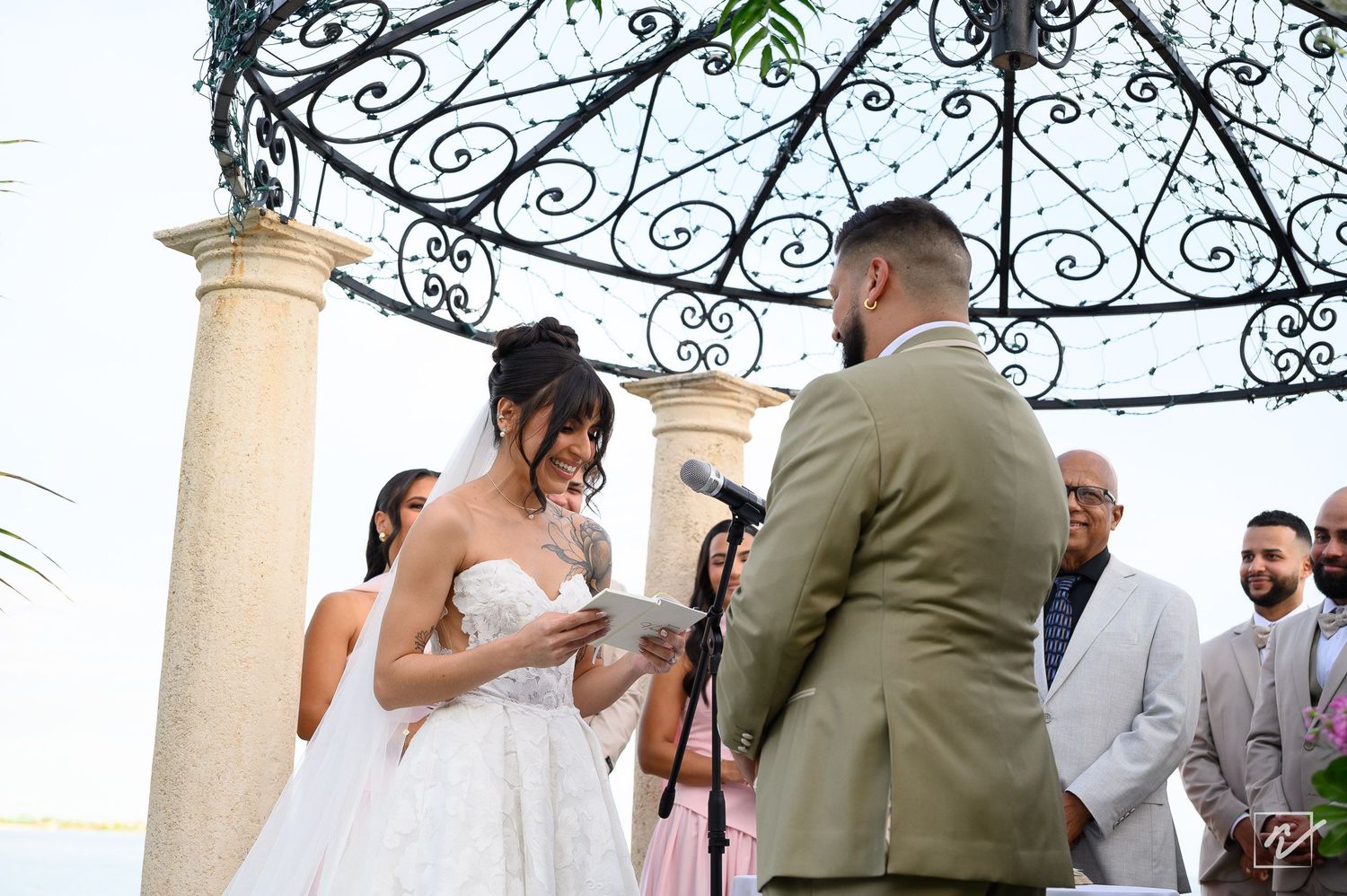 Bride in a white lace gown reads her vows to the groom in a tan suit under an ornate wrought iron gazebo as guests smile nearby.