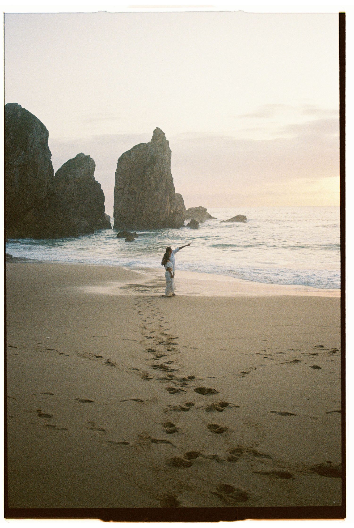 A film photograph of a couple on a misty beach at sunset with dramatic rock formations in Portugal