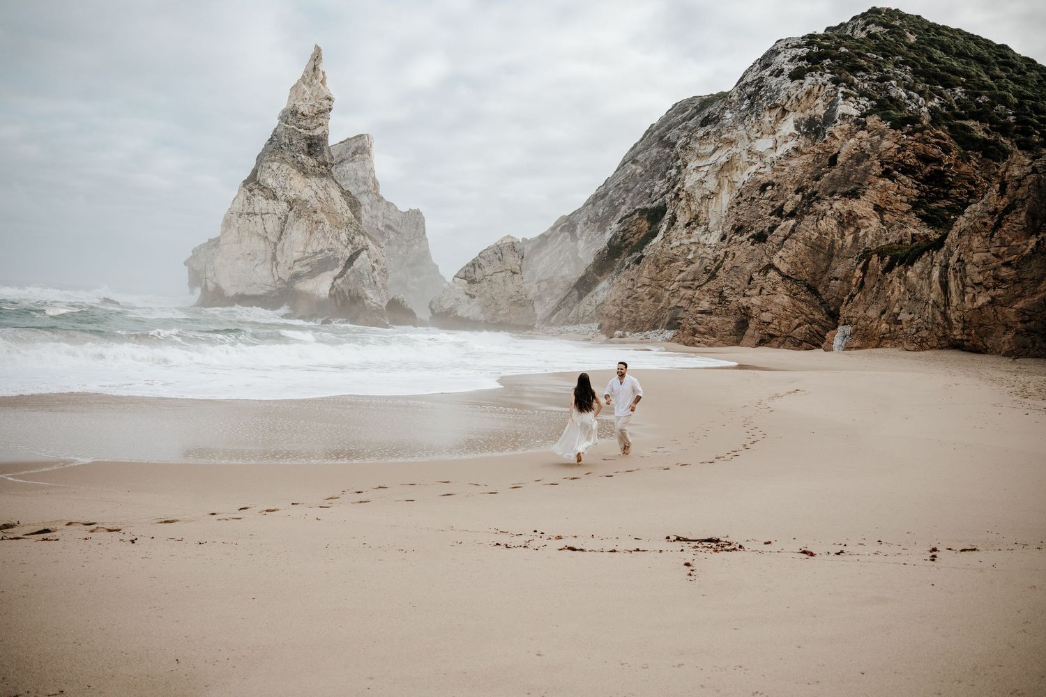 A couple walking on Praia da Ursa beach with dramatic rock formations rising from the sand