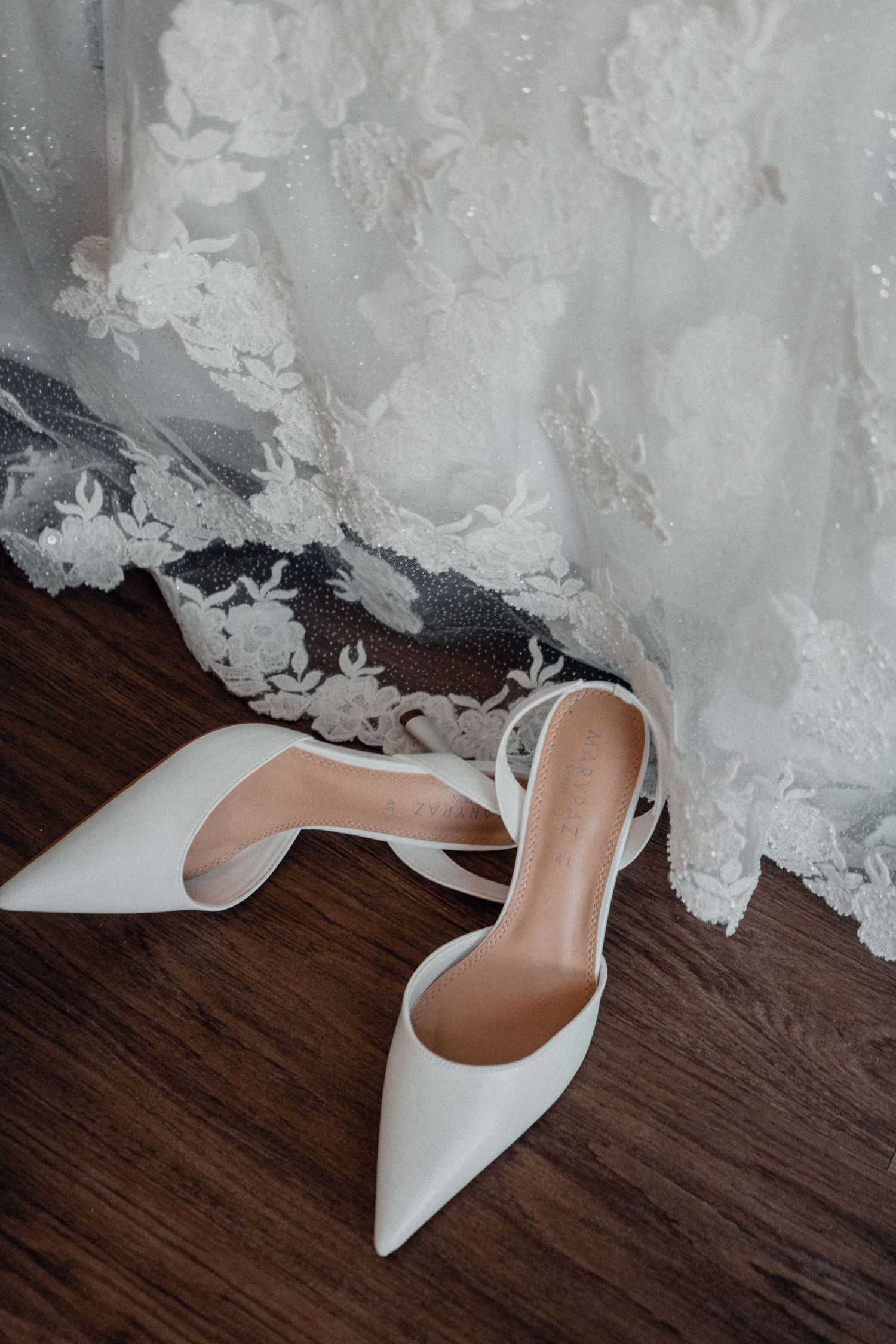 Elegant white bridal shoes on dark wooden flooring next to lace dress details.