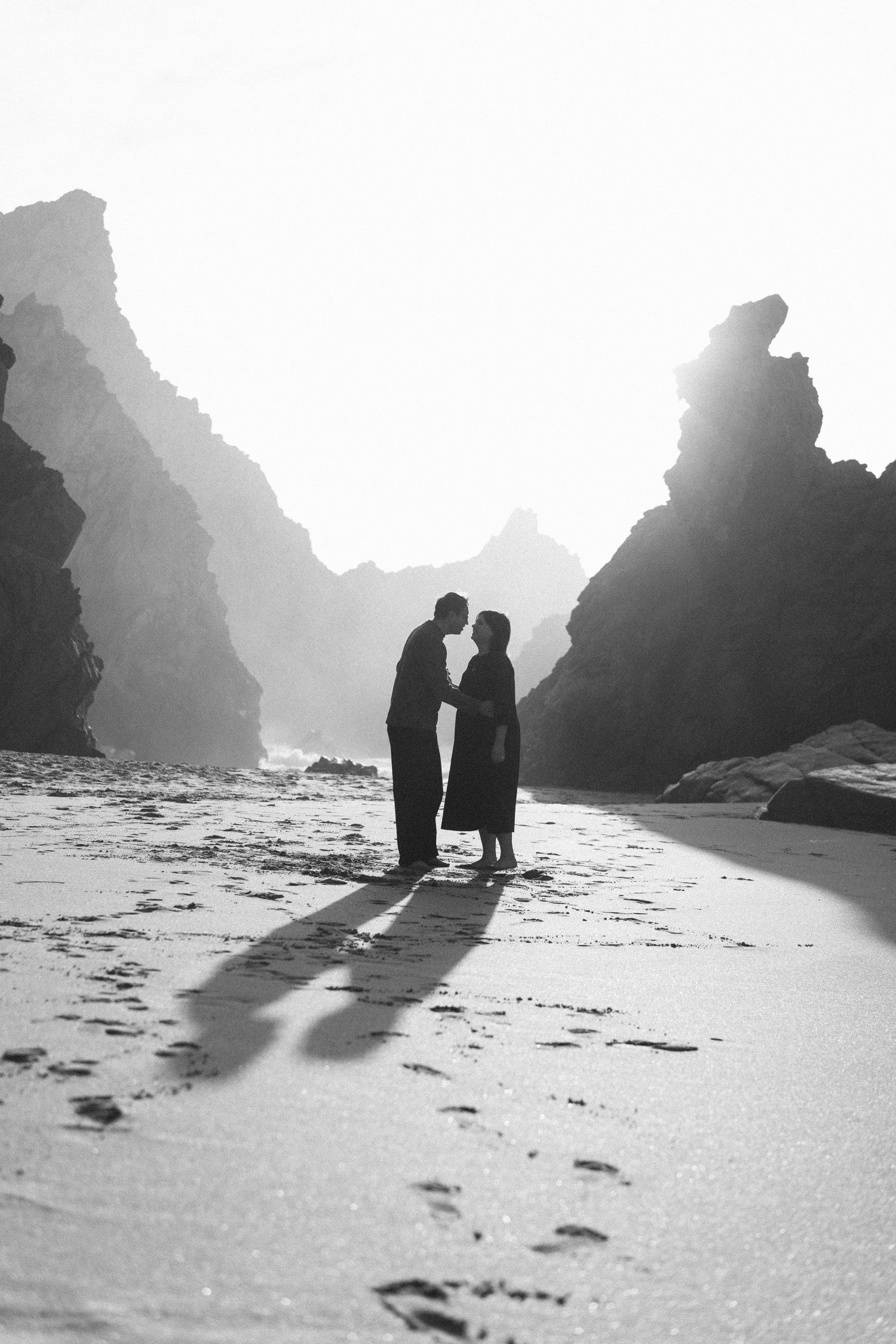 A back and white photo of a couple on Praia da Ursa with dramatic rock formations in the mist during their couples session in Portugal