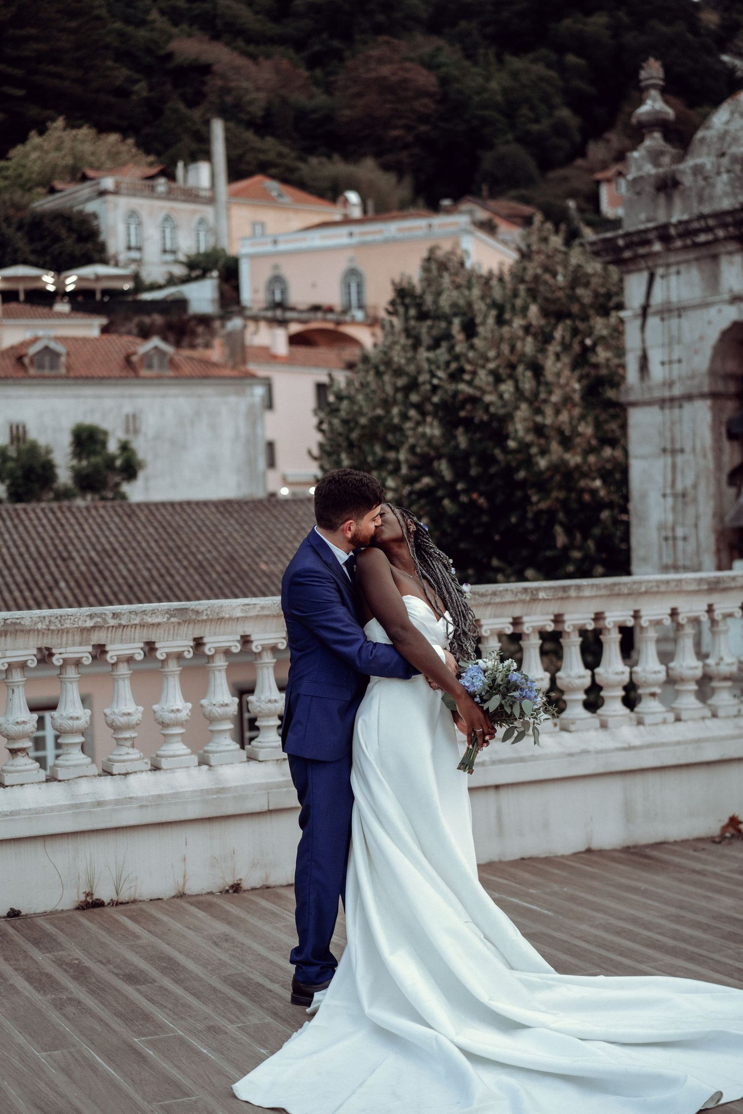 A couple in wedding attire embraces on a terrace with white balustrades and coastal buildings behind them.