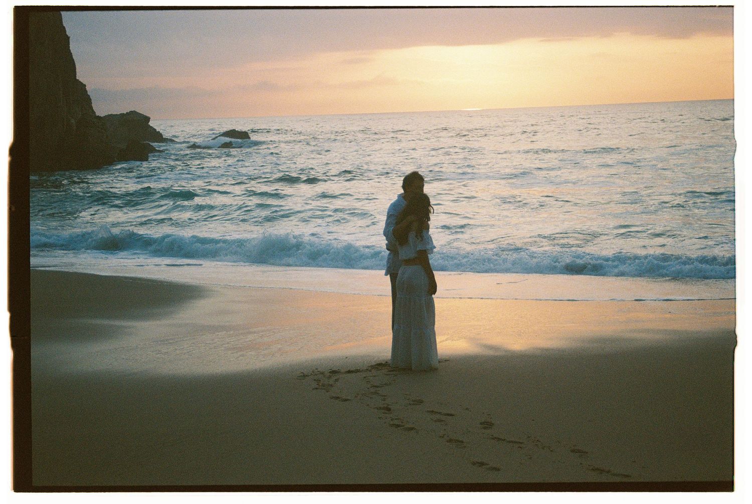 A film photograph of a couple standing on a beach at sunset with dramatic coastal rock formations in Portugal