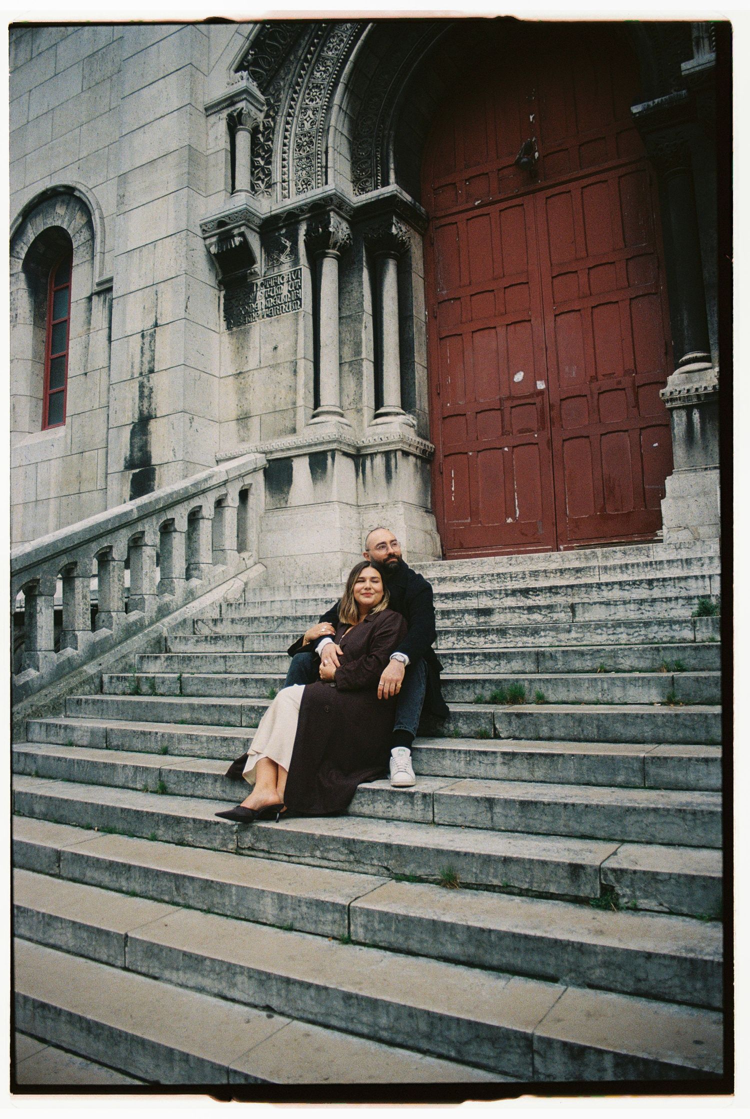 A film photograph of a couple sitting together on the grand steps of Sacré-Cœur at sunrise during a pre-wedding session in Montmartre, Paris