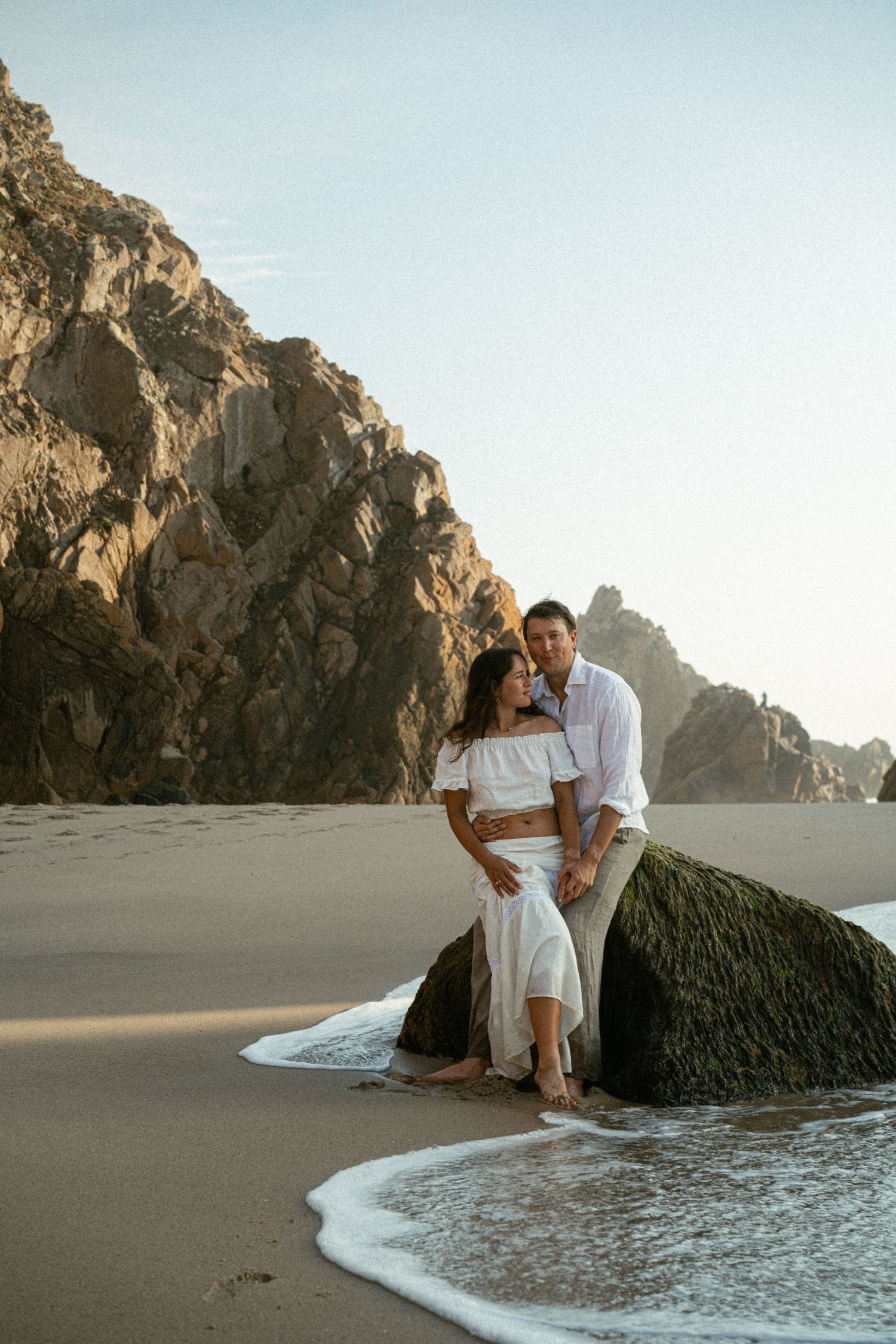 A couple sitting together on the rocks during a couple photoshoot on a beach in Portugal