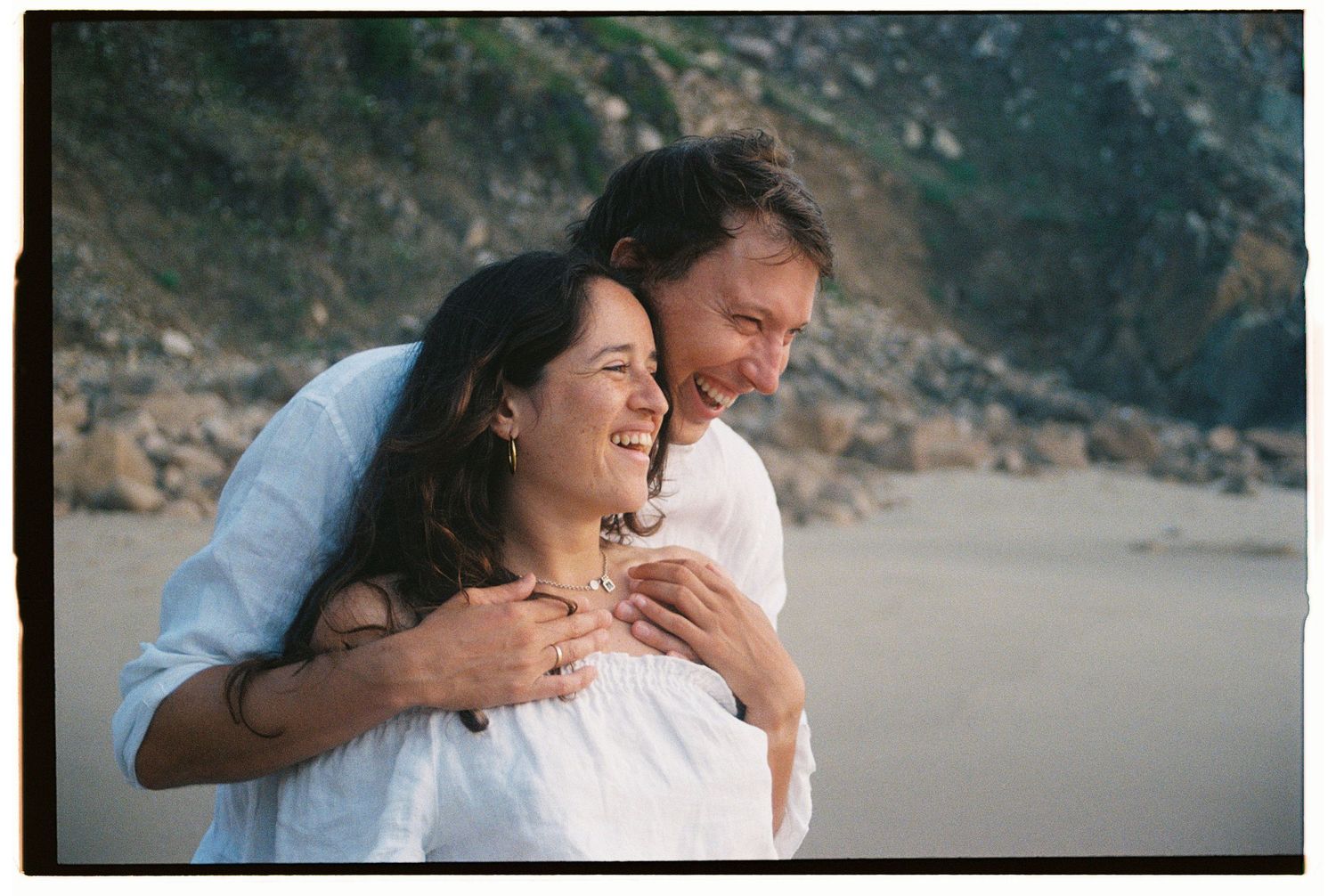 A film photograph of a couple laughing together on a rocky coastline during a couples session on a beach in Portugal