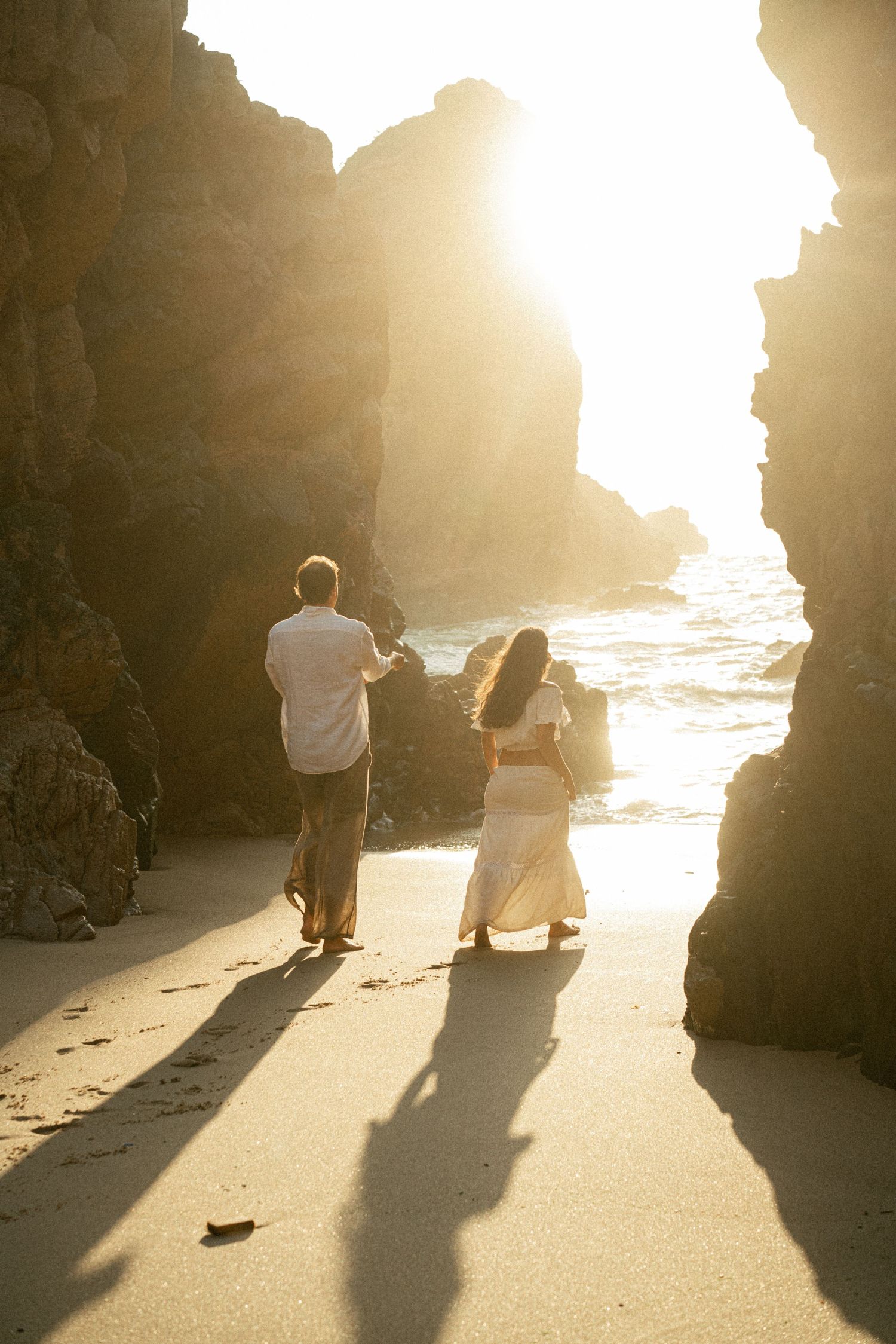 A couple walking together on Praia da Ursa at golden hour with dramatic rock formations behind them