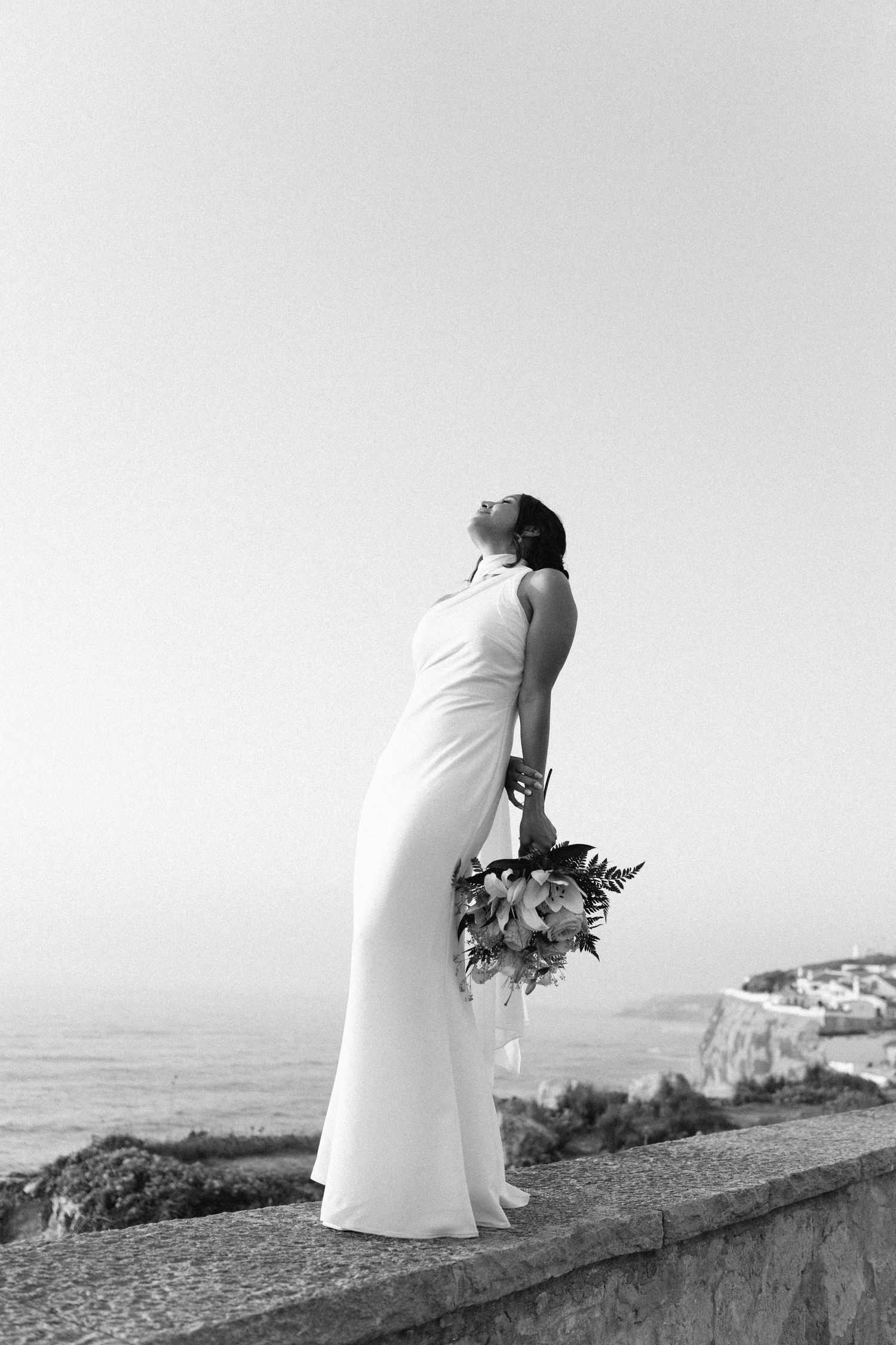 A bride in a sleek white dress holding a bouquet while standing on a coastal cliff.