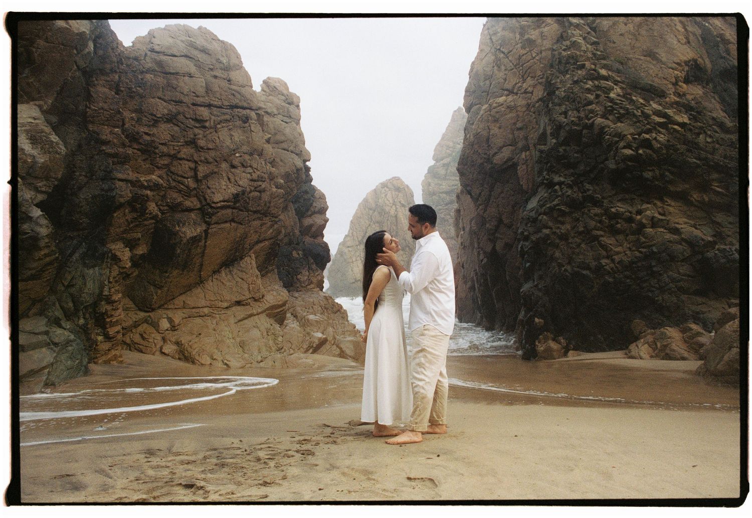 A film photograph of a couple standing between dramatic rocky cliffs on a beach in Portugal