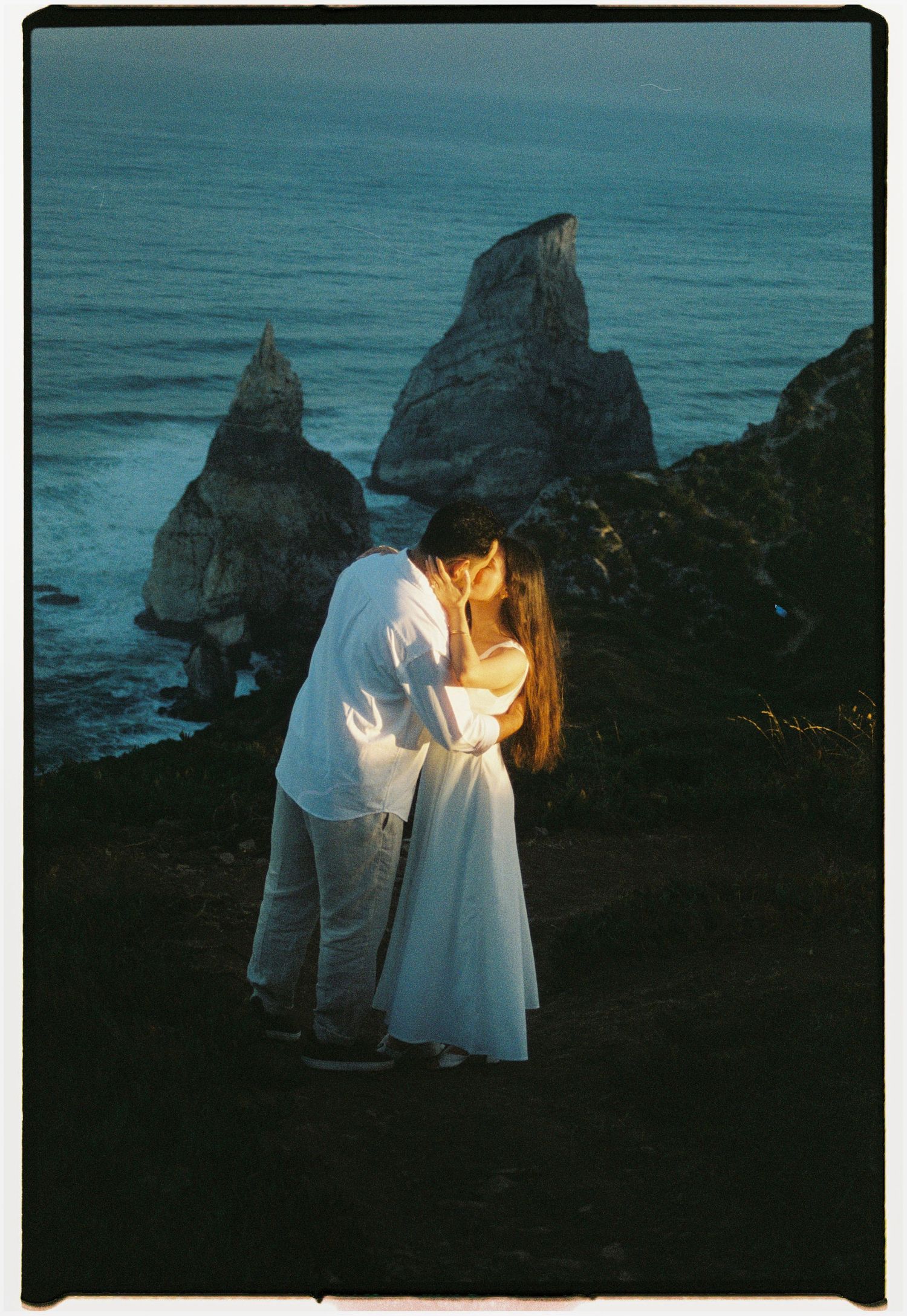 A film photograph of a couple embracing on Praia da Ursa at dusk with dramatic rock formations behind them, Portugal